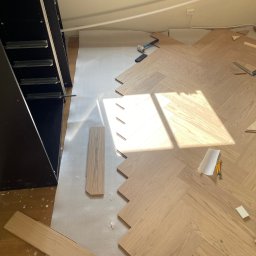 SEB&SON - Układanie jasnego parkietu w jodełkę francuską, widoczne narzędzia i fragment szafy wnękowej. Częściowo ułożona podłoga w słonecznym wnętrzu.