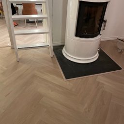 Maja Kienitz-Sierakowska SEB&SON - Jasny parkiet ułożony w jodełkę, biały piec kaflowy na czarnej podstawie i biała drabina w tle. Widoczne detale wykończenia wnętrza.