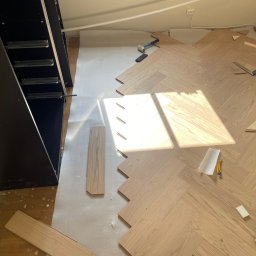 Układanie jasnego parkietu w jodełkę francuską, widoczna część szafy w zabudowie, narzędzia pracy i fragmenty desek. Podłoże zabezpieczone folią.