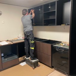 Maja Kienitz-Sierakowska SEB&SON - Monterażysta na podeście montuje górne szafki kuchenne w surowym wnętrzu. Widoczne narzędzia, elementy zabudowy i kartonowe zabezpieczenie podłogi.