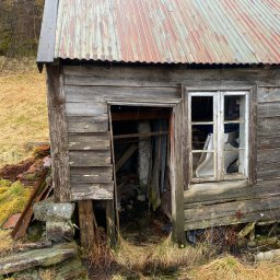 Maja Kienitz-Sierakowska SEB&SON - Zrujnowana, drewniana szopa z blachą falistą na dachu, z wybitym oknem i otwartymi drzwiami, otoczona trawą i kamieniami. Widoczne ślady zaniedbania i erozji.