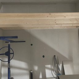 Maja Kienitz-Sierakowska SEB&SON - Wnętrze w trakcie prac wykończeniowych: surowe drewno antresoli, płyty gipsowo-kartonowe na ścianach z widocznymi otworami i narzędzie do montażu płyt.