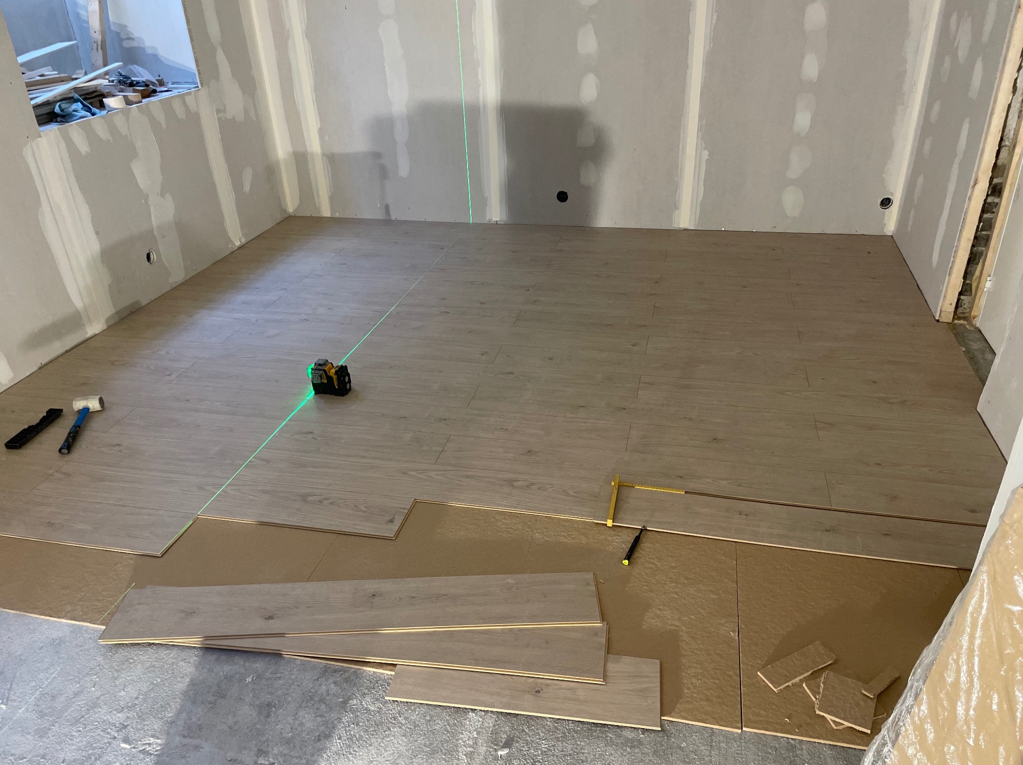 Układanie paneli podłogowych w pomieszczeniu z surowymi ścianami. Widoczny laser krzyżowy, narzędzia i panele w trakcie montażu. Jasne panele, surowy stan wykończenia.