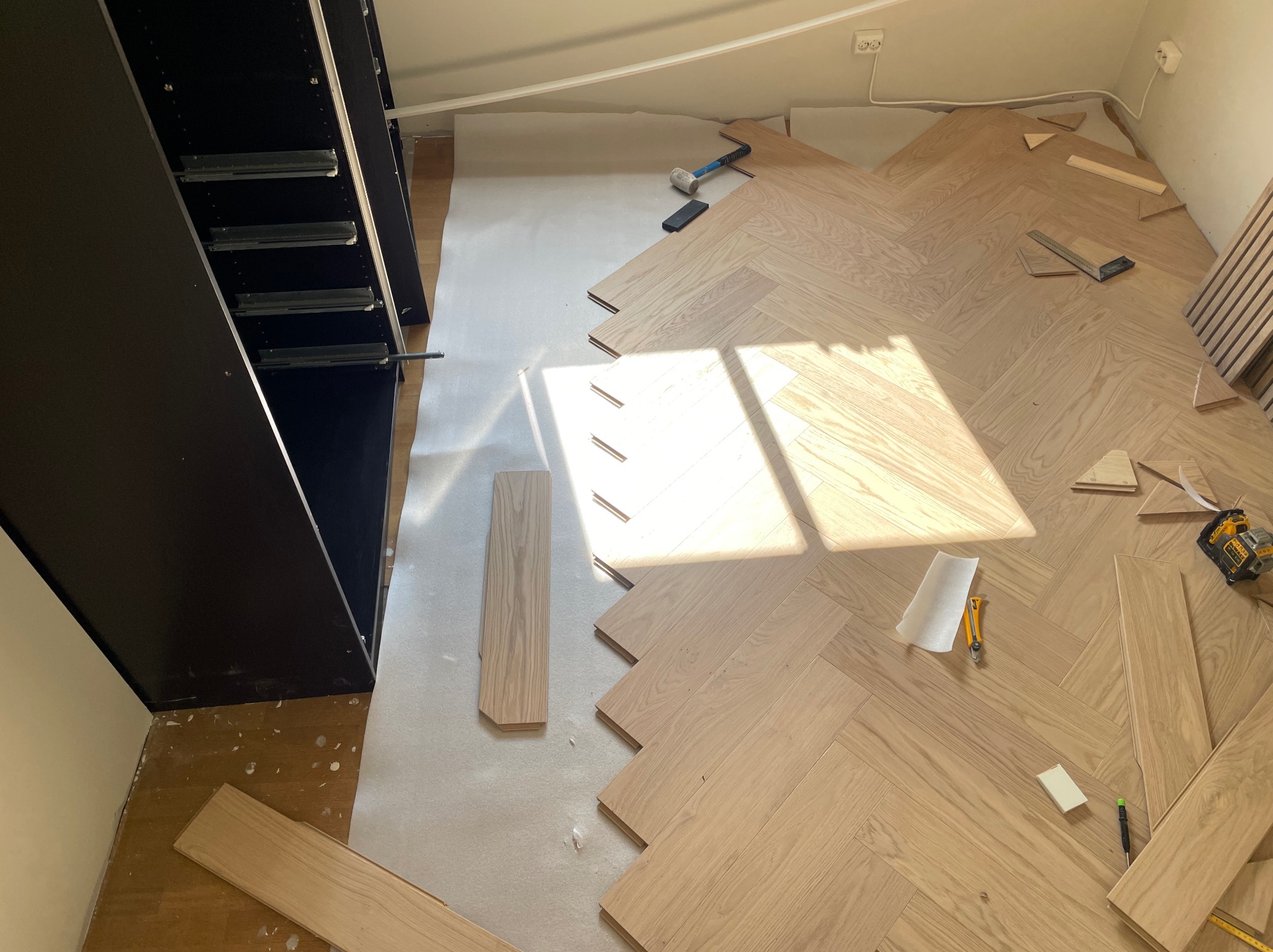 Układanie jasnego parkietu w jodełkę francuską, widoczne narzędzia i fragment szafy wnękowej. Częściowo ułożona podłoga w słonecznym wnętrzu.