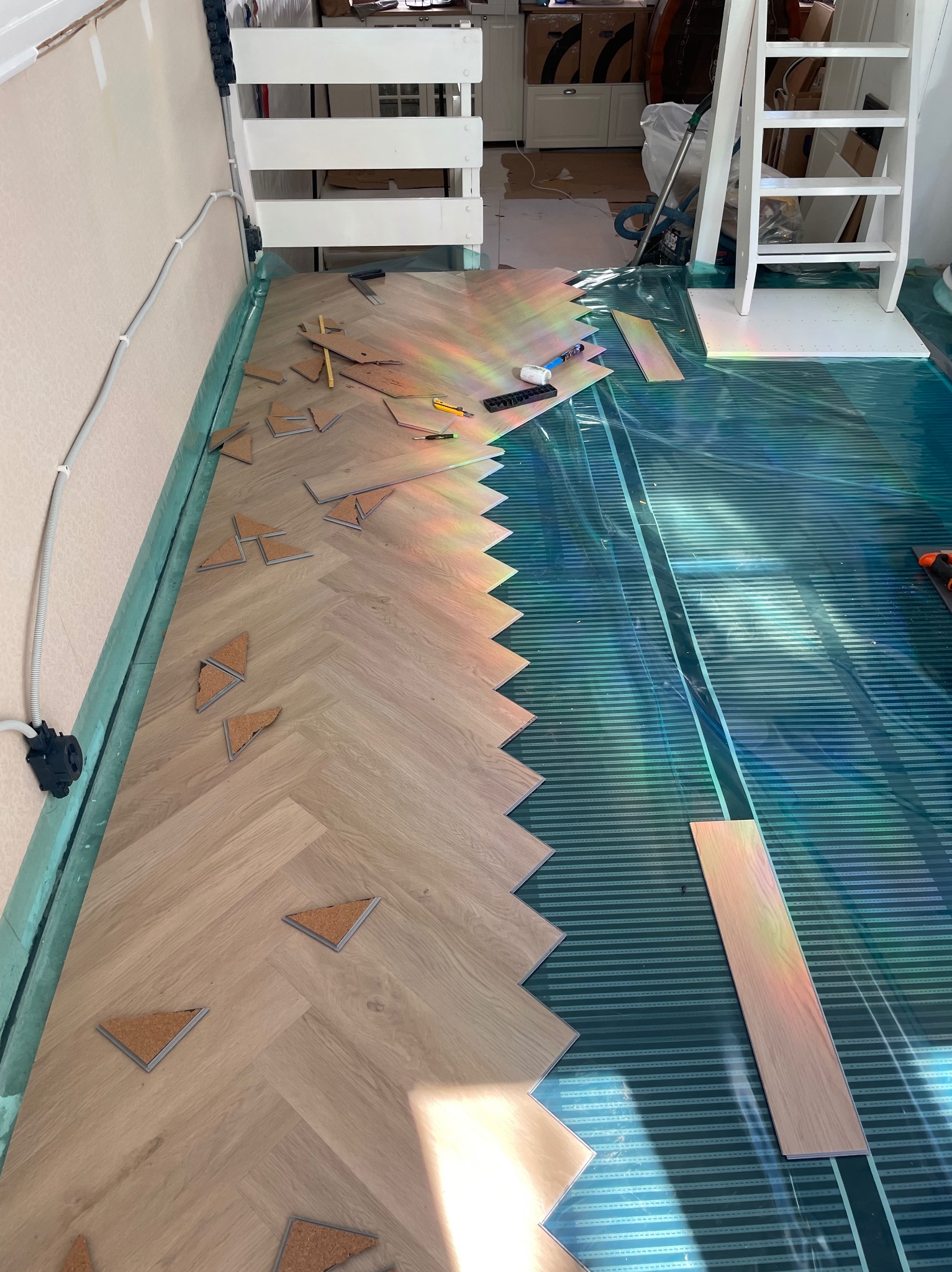 Układanie parkietu w jodełkę na folii grzewczej, widoczne narzędzia i fragment schodów. Częściowo ułożona podłoga z drewnianych paneli, detale montażu.