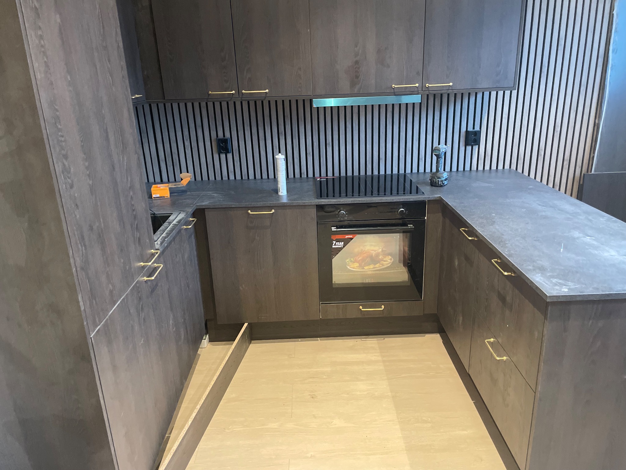 Ciemna, nowoczesna kuchnia z drewnianymi frontami i złotymi uchwytami. Widoczny piekarnik, płyta indukcyjna i pionowe panele dekoracyjne na ścianie. W tle narzędzia.