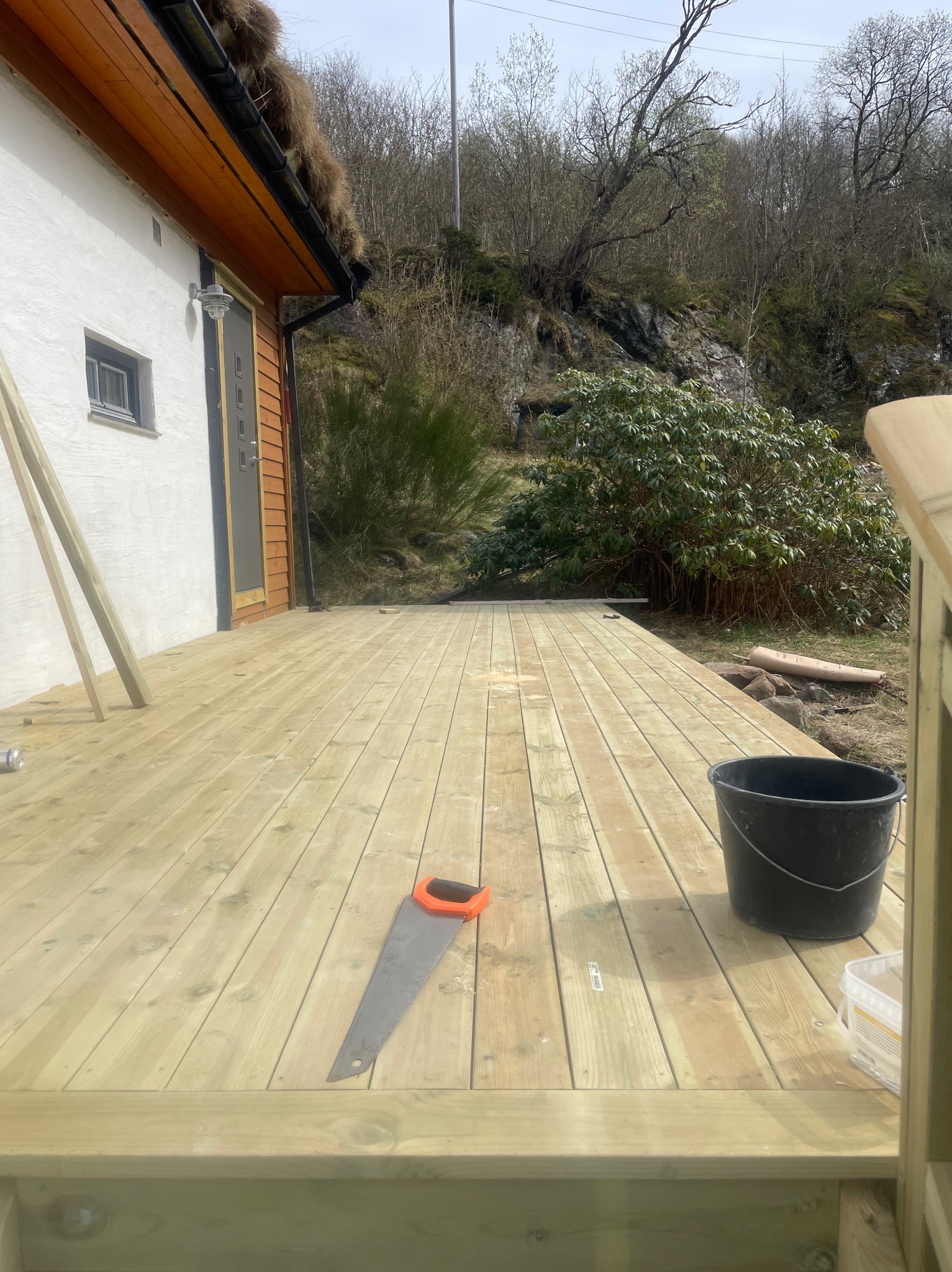 Jasny, drewniany taras w trakcie budowy przy domu z dachem porośniętym trawą. Piła ręczna i wiadro na deskach. Tło stanowią drzewa i skały.