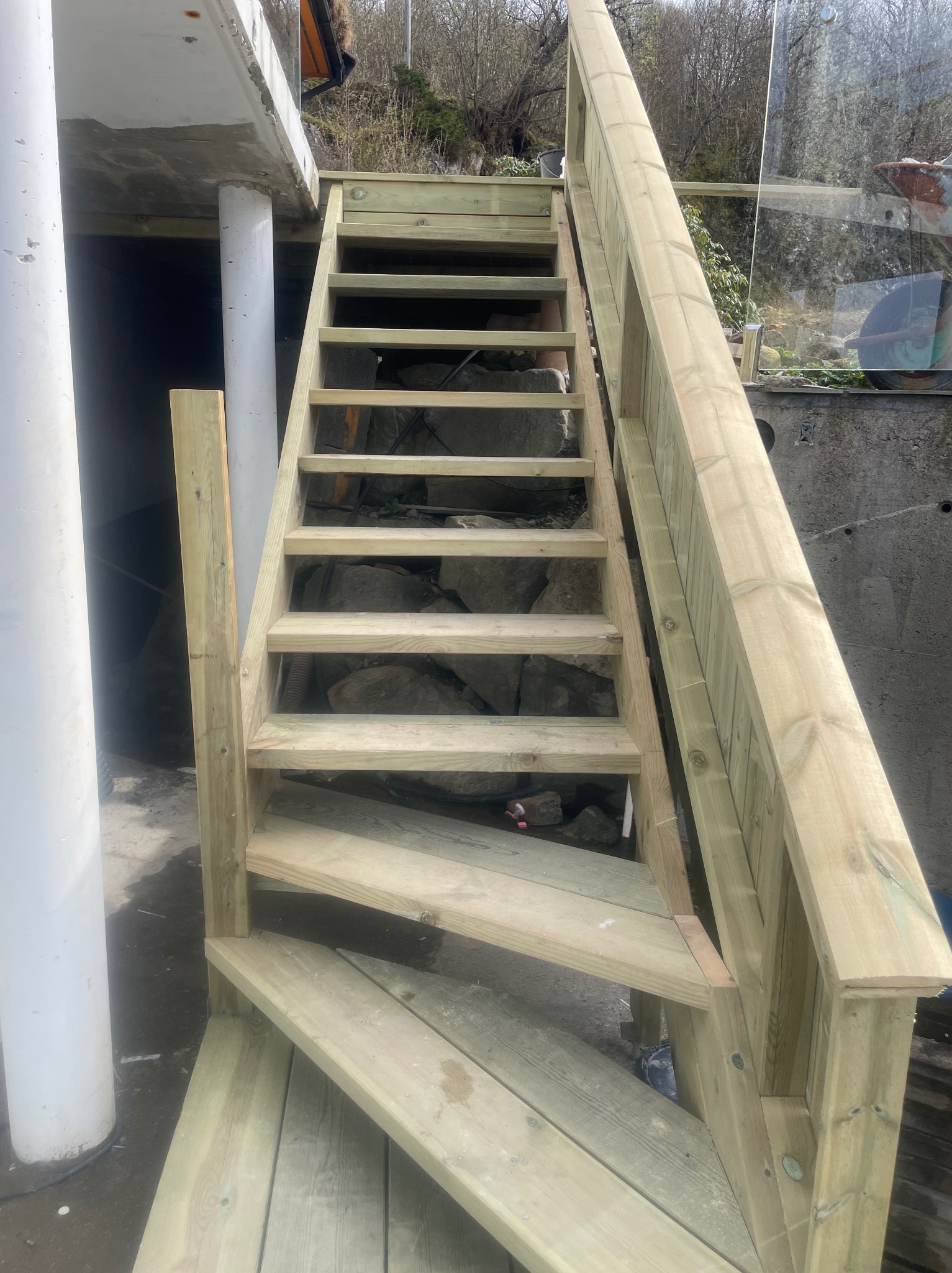 Zewnętrzne, surowe schody drewniane z jasnego drewna prowadzące do góry, z widocznymi kamieniami pod spodem. Minimalistyczny design, naturalne otoczenie.