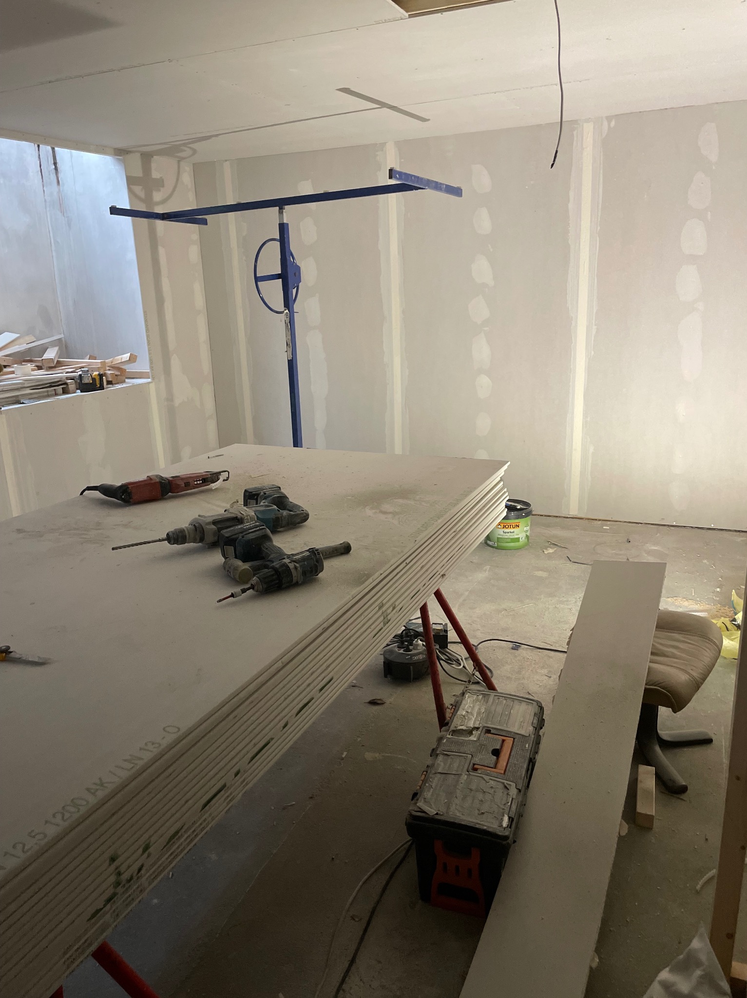 Wnętrze w trakcie wykończenia: płyty gipsowo-kartonowe na stojaku, narzędzia (wiertarki, szlifierka), podnośnik do płyt, puszka z masą szpachlową. Widoczne ślady szpachlowania na ścianach.