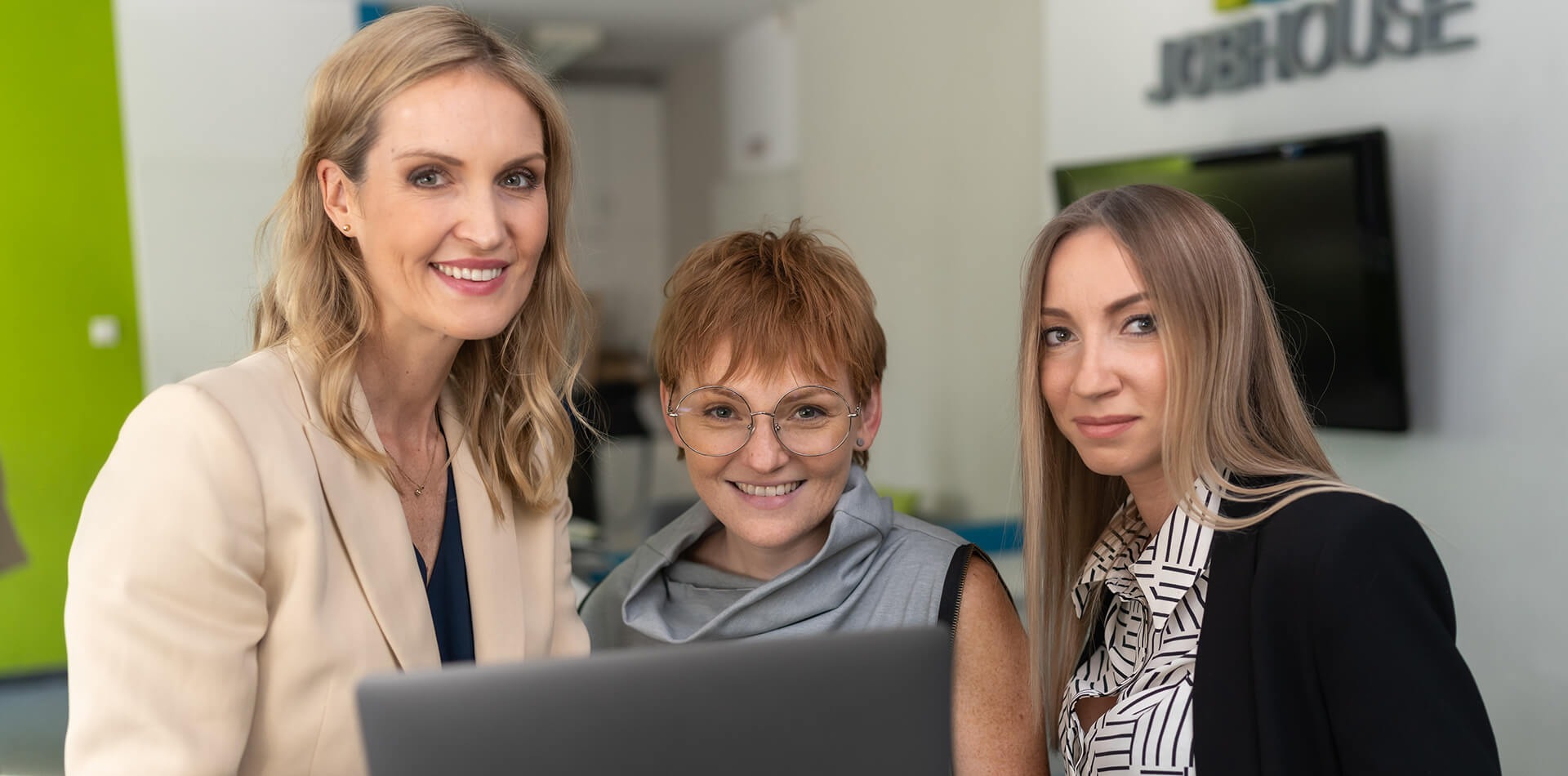 Trzy uśmiechnięte kobiety w biurze Jobhouse, patrzące w obiektyw, z laptopem w kadrze. Profesjonalny zespół agencji rekrutacyjnej w jasnym wnętrzu.
