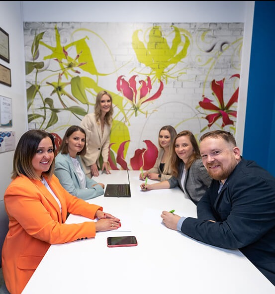 Zespół agencji rekrutacyjnej podczas spotkania w biurze z muralem kwiatowym. Widoczne osoby przy stole, laptop i telefon. Kobieta w pomarańczowej marynarce na pierwszym planie.