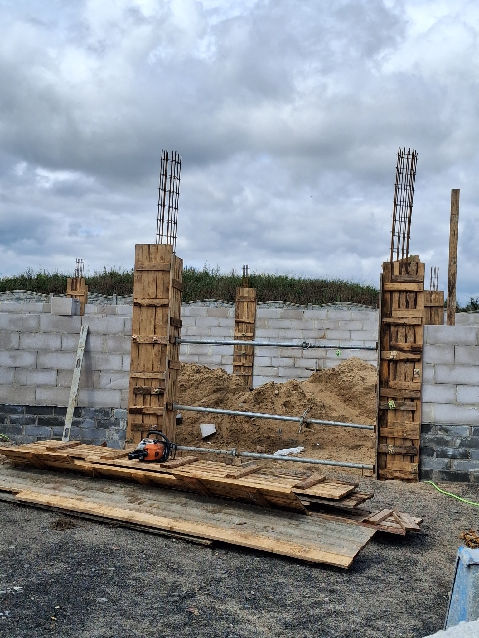 Budowa fundamentów: drewniane szalunki z wystającą stalą zbrojeniową, piasek w środku, piła łańcuchowa na deskach, bloczki betonowe w tle, pochmurne niebo.