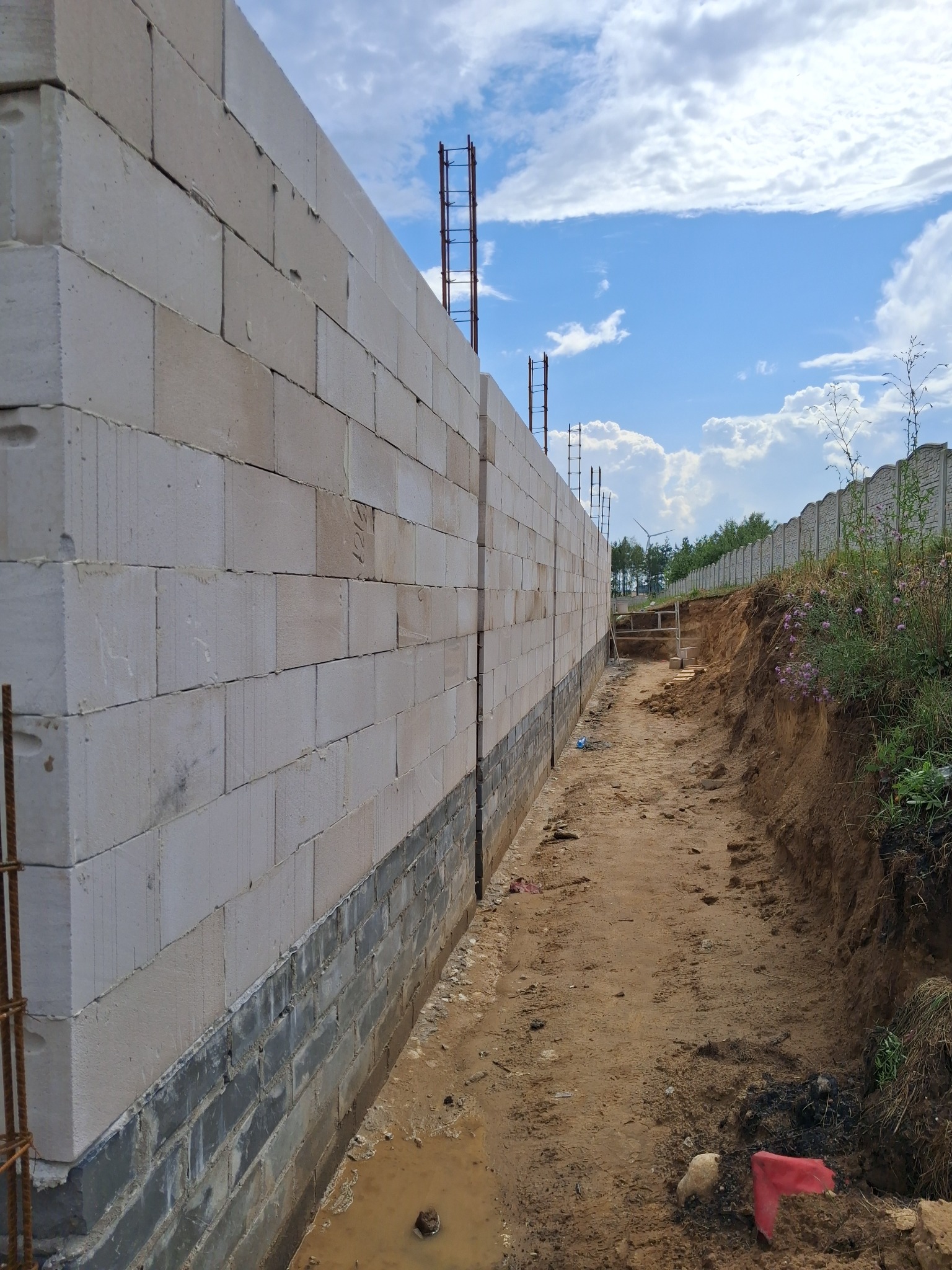 Ściana z białych bloczków, częściowo podmurowana szarymi cegłami, z widocznym wykopem ziemnym wzdłuż niej. W tle błękitne niebo z chmurami i fragment ogrodzenia.