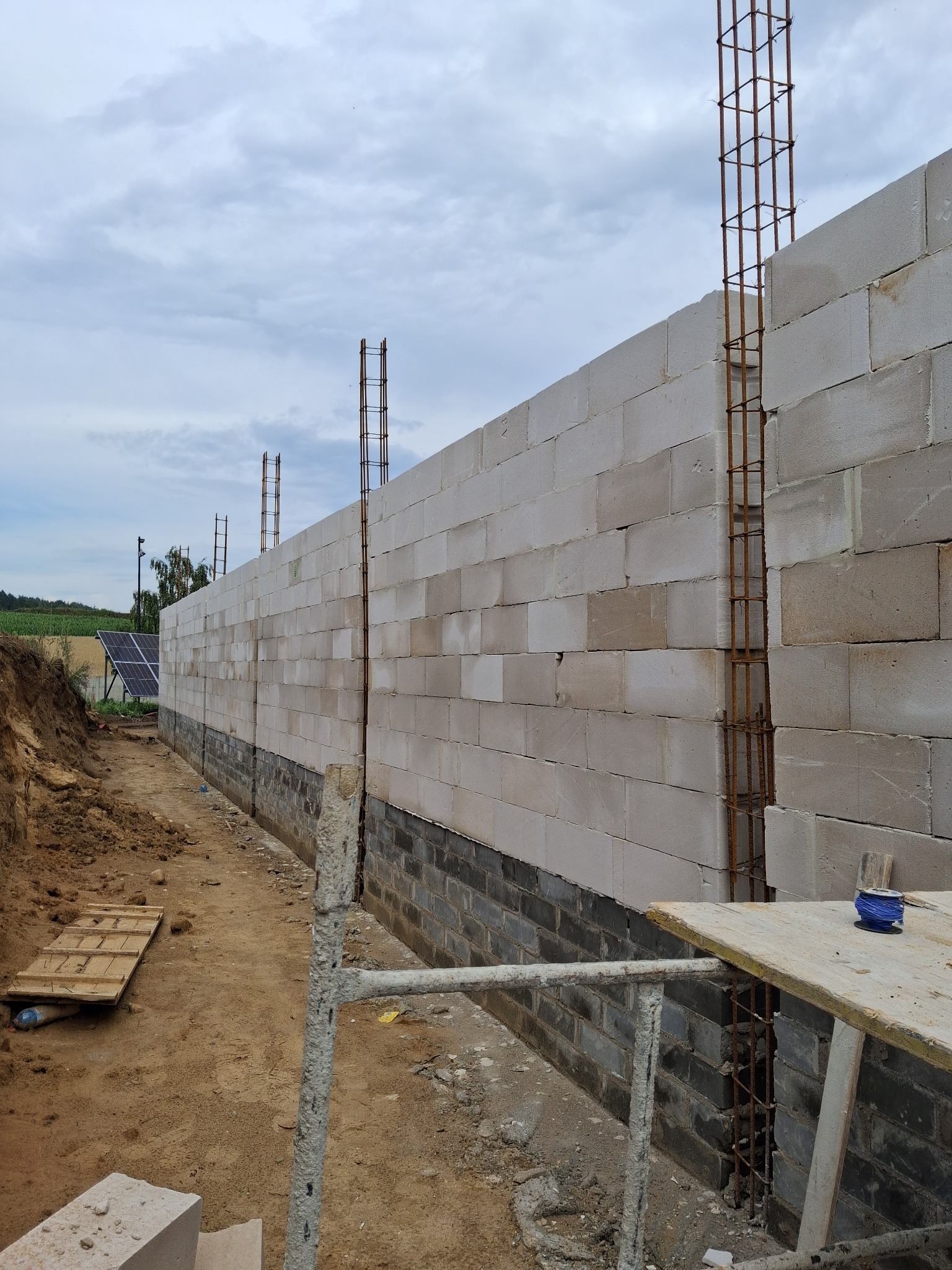Wznoszenie ściany fundamentowej z bloczków betonowych z widocznym zbrojeniem pionowym. Na pierwszym planie fragment rusztowania i wykop.