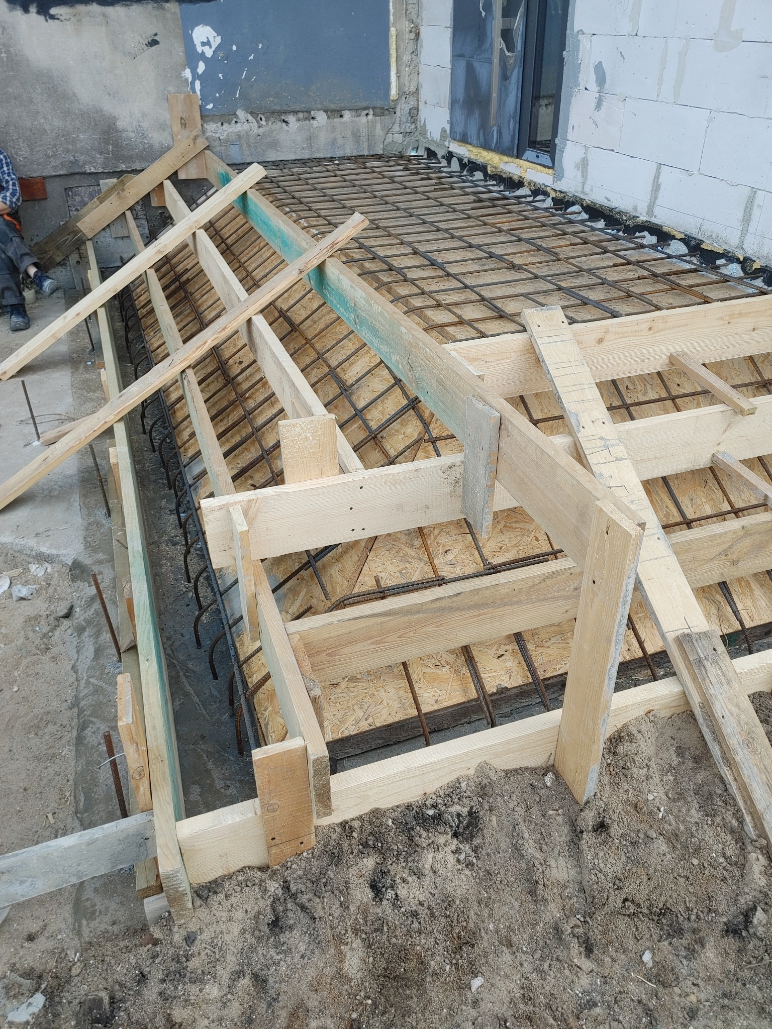 Zbrojenie fundamentów pod budynek z drewnianym szalunkiem, widoczne pręty zbrojeniowe i deski szalunkowe. Etap przygotowawczy do wylewania betonu.