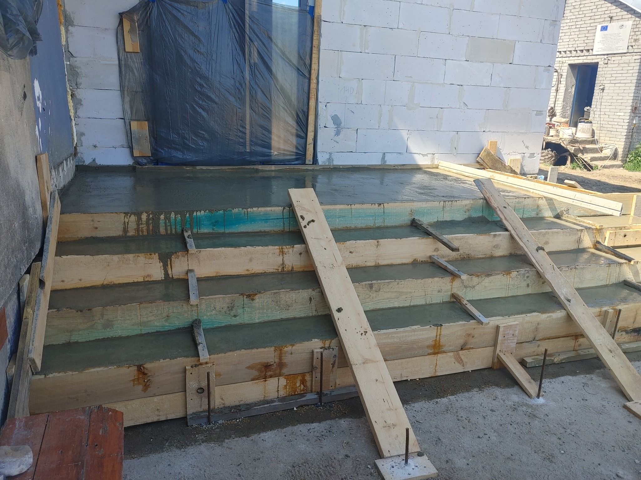 Betonowe schody w trakcie budowy, drewniane szalunki, widoczne pręty zbrojeniowe, budynek w tle z częściowo zasłoniętym wejściem folią.