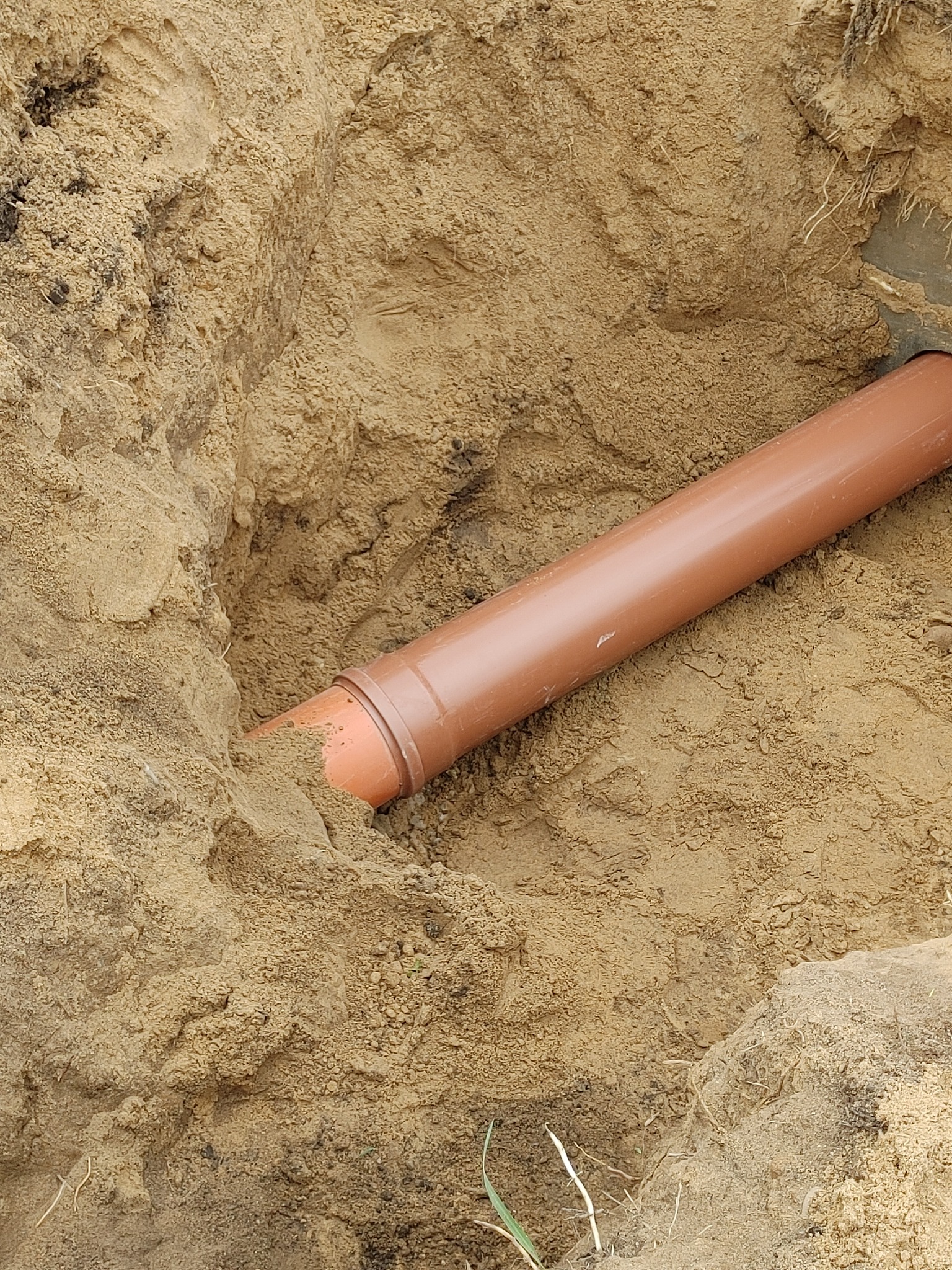 Brązowa rura kanalizacyjna ułożona w wykopie wypełnionym piaskiem. Widoczne ślady po wykopie i nierówności terenu. Fragment trawy w dolnej części kadru.