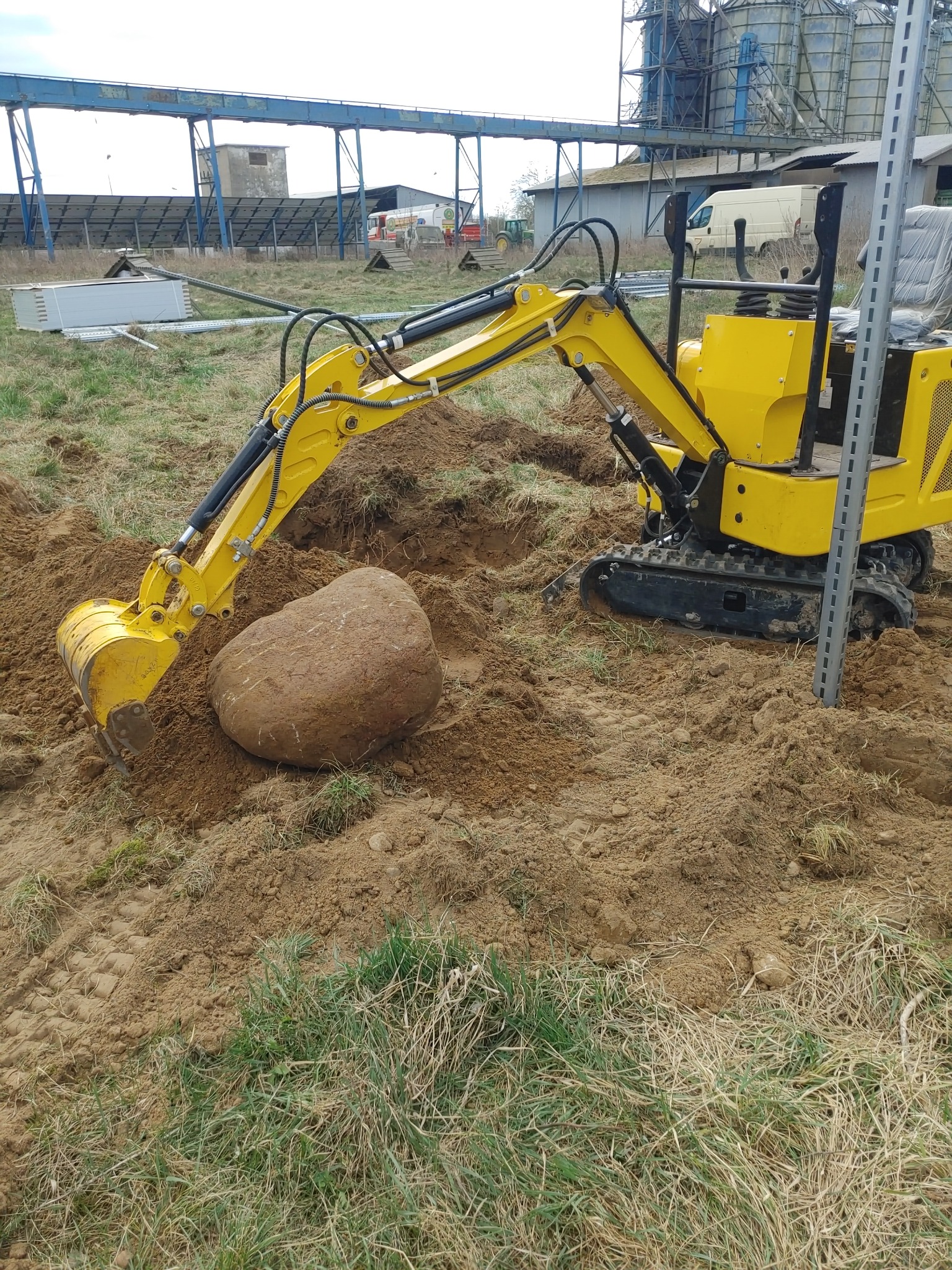 Żółta mini koparka z łyżką podnosi duży kamień na ziemi. W tle budynki gospodarcze i konstrukcja stalowa.