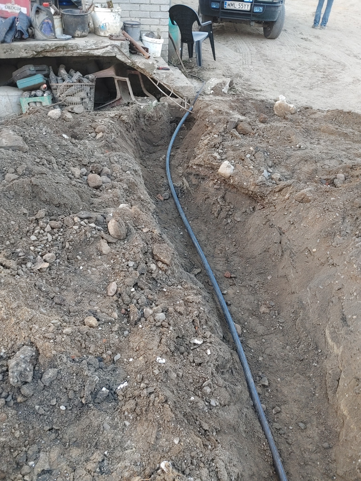 Wąski wykop w ziemi z ułożonym kablem, widoczne narzędzia i fragment samochodu w tle. Prace ziemne w trakcie realizacji.