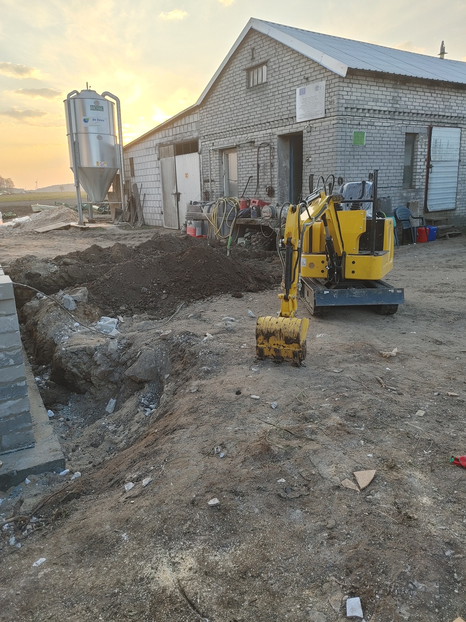Żółta mini koparka przy wykopie fundamentowym na placu budowy, w tle budynek gospodarczy i silos. Prace ziemne w trakcie realizacji, widoczne narzędzia i sprzęt.