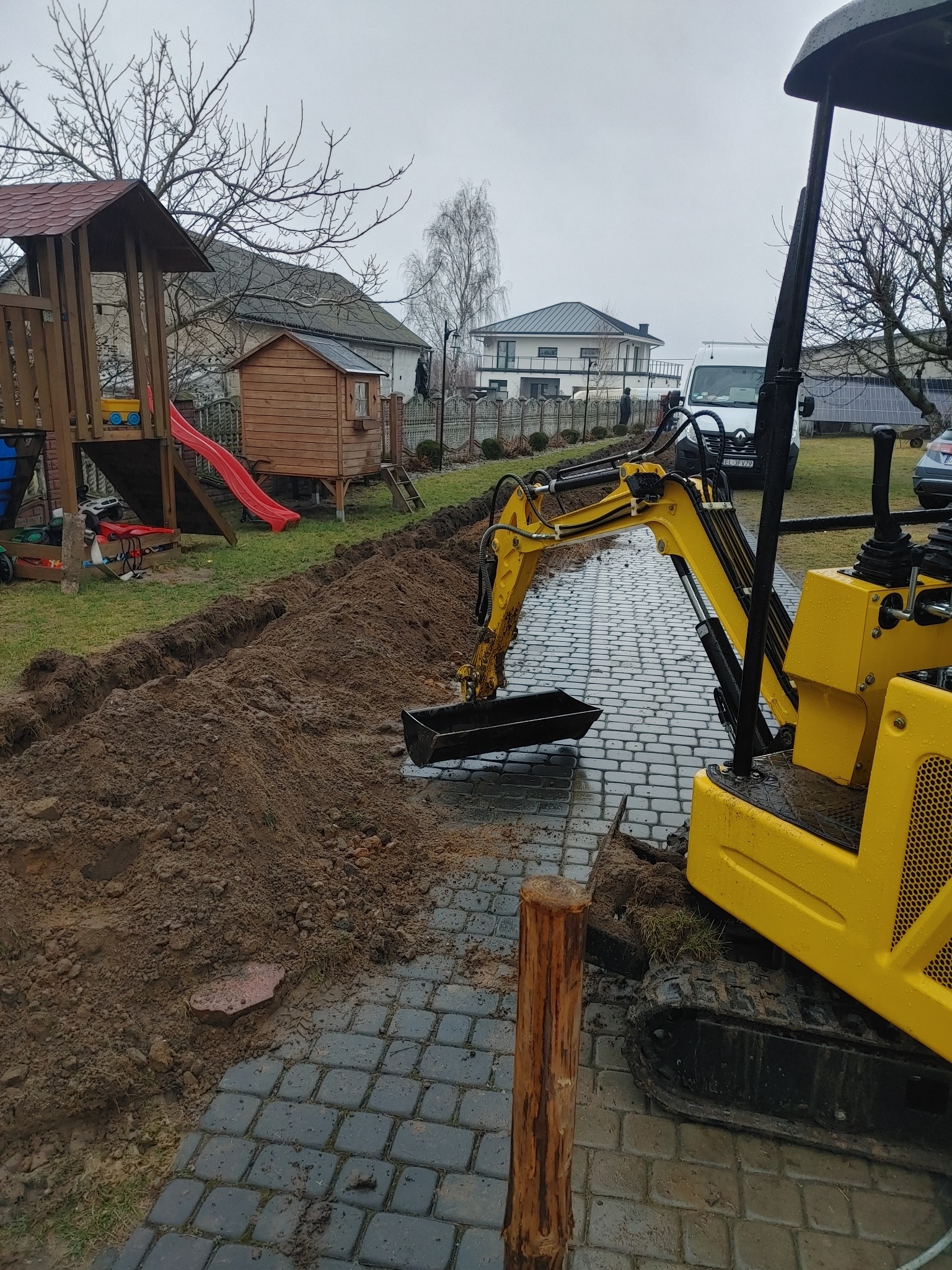 Żółta mini koparka na kostce brukowej, wykonująca wykop w ziemi. W tle plac zabaw, dom i samochody. Widoczne ślady błota na kostce i maszynie.