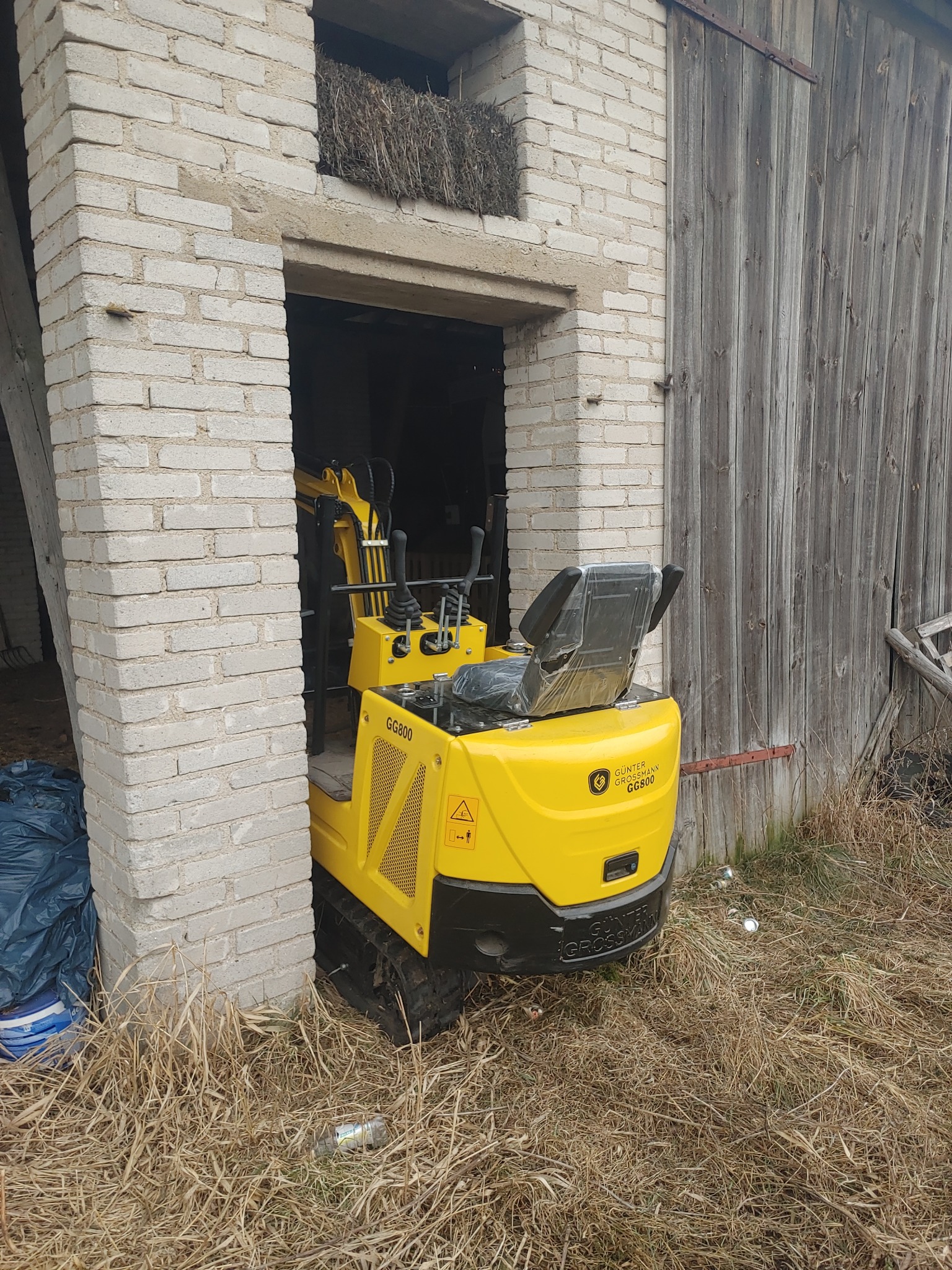 Żółta mini koparka Gunter Grossmann GG800 w otworze starego, ceglanego budynku gospodarczego z drewnianymi drzwiami. W tle sterta siana.