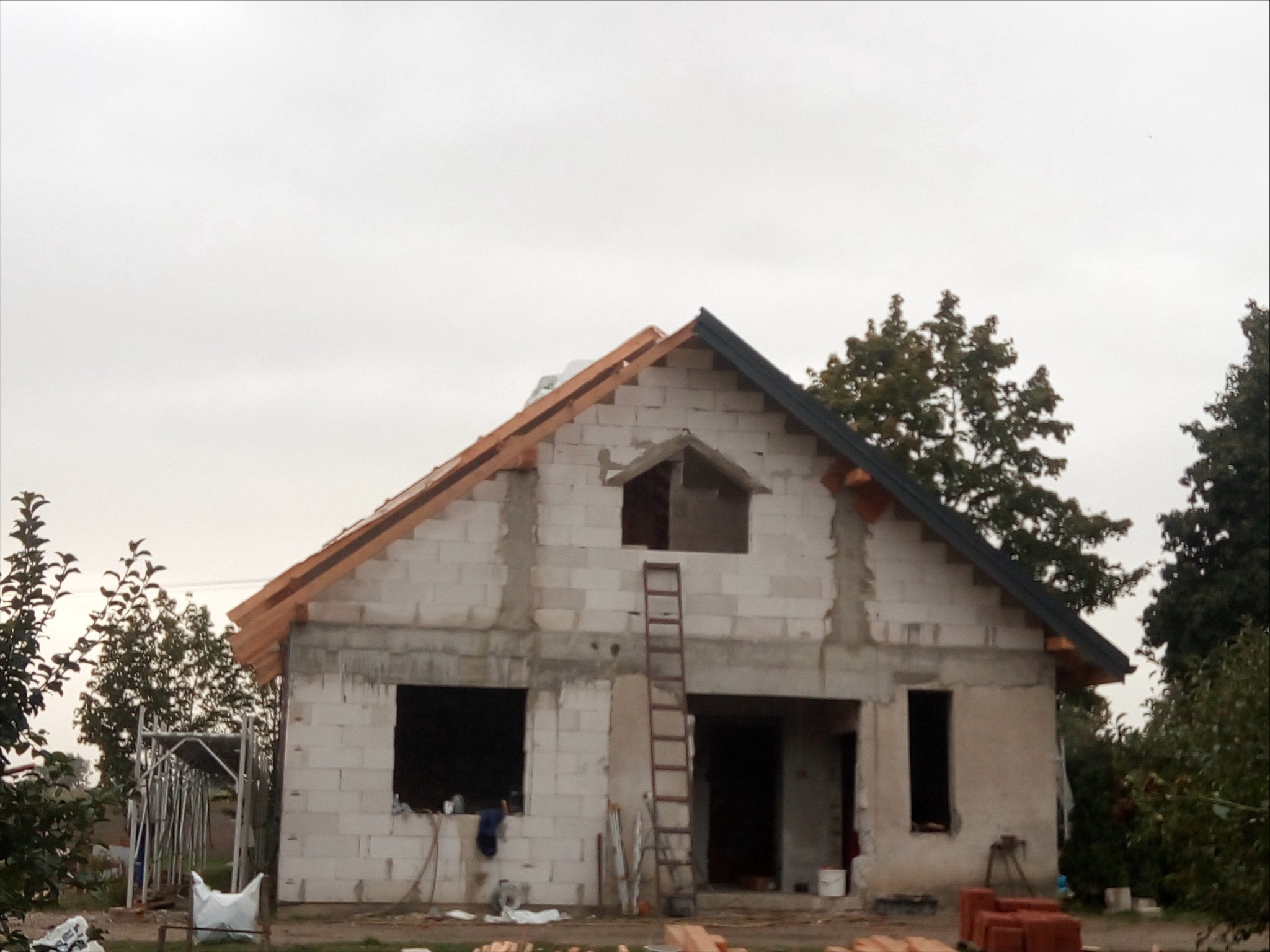 Surowy stan domu z białego bloczka, z częściowo ukończonym dachem i drewnianą konstrukcją więźby. Na tle pochmurne niebo i zieleń drzew. Widoczna drabina oparta o ścianę.