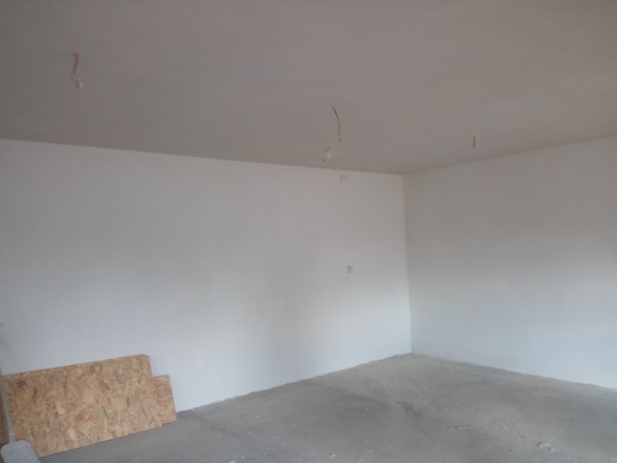 Surowe wnętrze w trakcie wykańczania: białe ściany, szara podłoga, wystające kable z sufitu i płyta OSB oparta o ścianę. Widok na róg pomieszczenia.