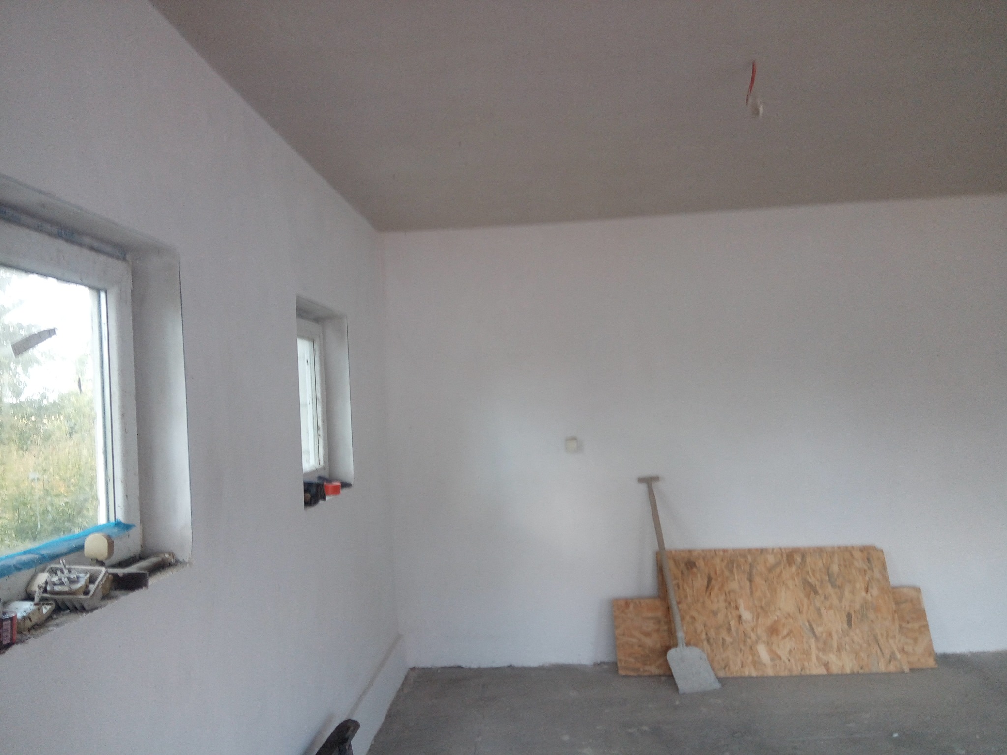 Wnętrze w trakcie prac wykończeniowych: świeżo otynkowane ściany, okna z narzędziami, płyty OSB i łopata przy ścianie. Widoczny surowy stan podłogi.