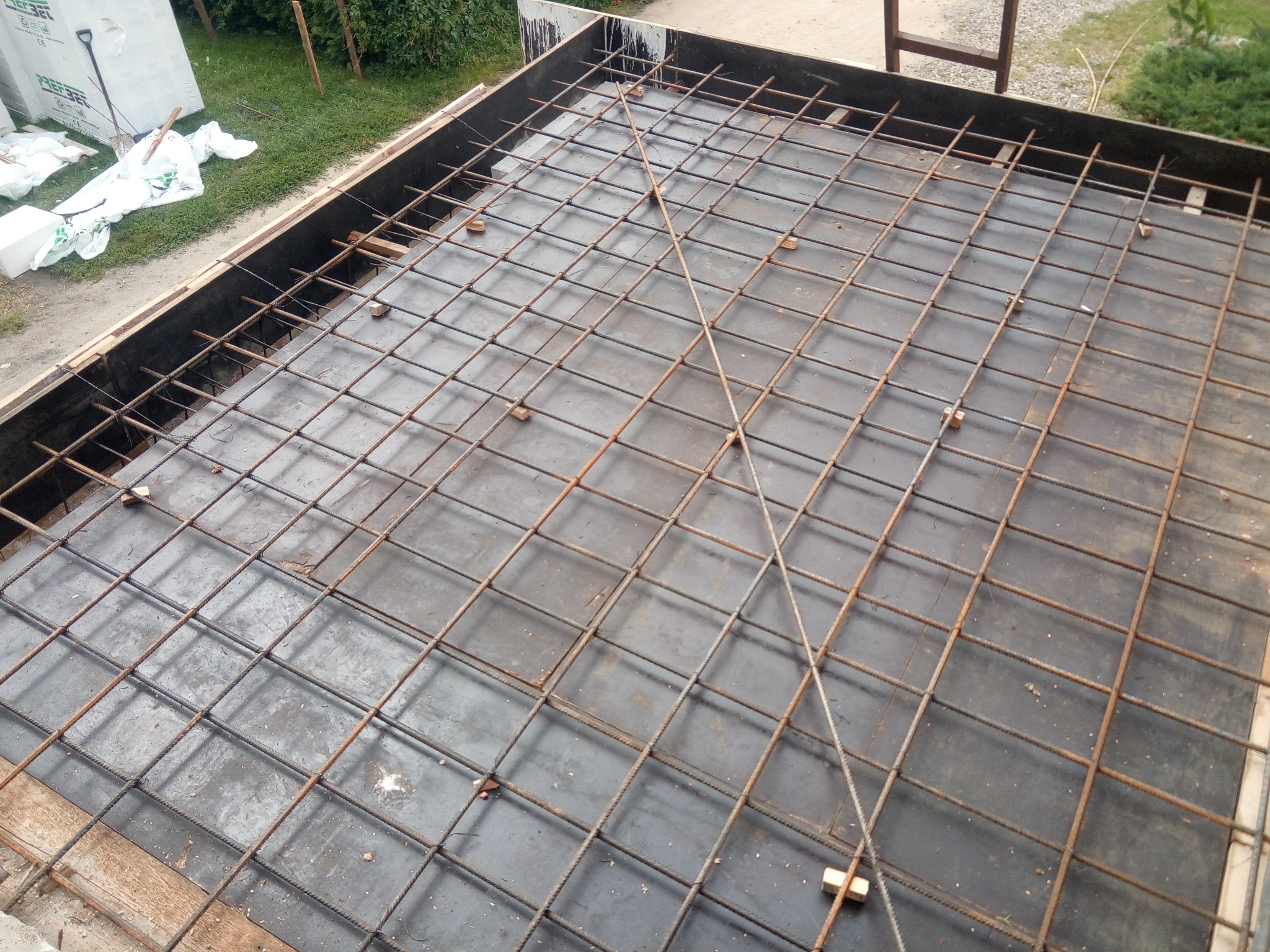 Zbrojenie fundamentu w szalunku, widok z góry. Siatka z prętów stalowych ułożona na folii, przygotowana do zalania betonem. W tle widoczne narzędzia i materiały budowlane.