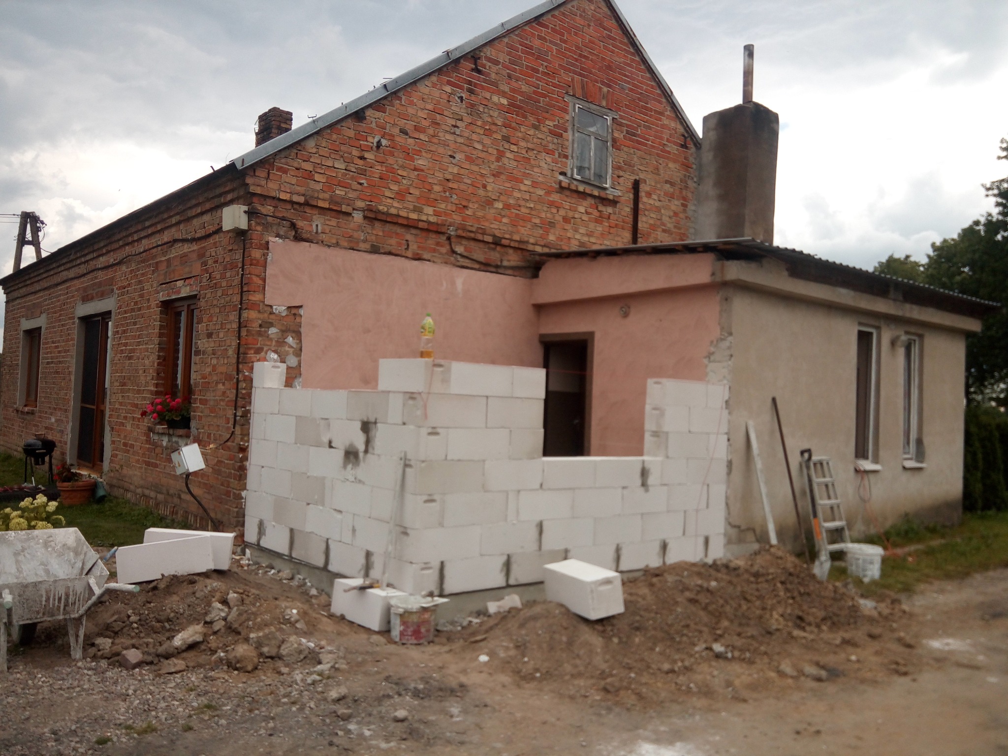 Budowa domu z czerwonej cegły: wznoszenie ściany z białych bloczków, widoczne narzędzia i gruz. Etap prac murarskich przy dobudowie.