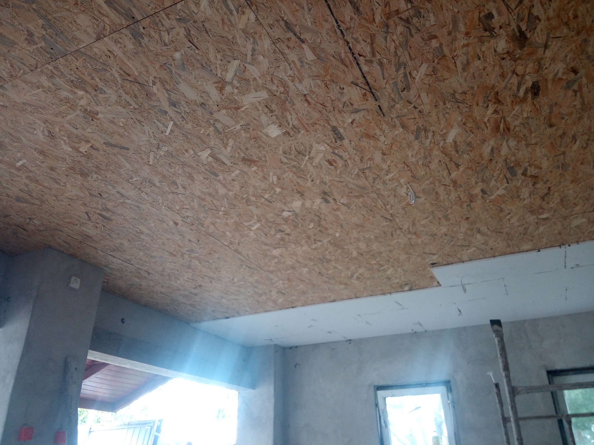 Wnętrze w trakcie budowy: sufit wykończony płytami OSB, częściowo pokryty białymi panelami. Widoczne fragmenty ścian i okien, drabina oparta o ścianę. Surowy stan wykończenia.