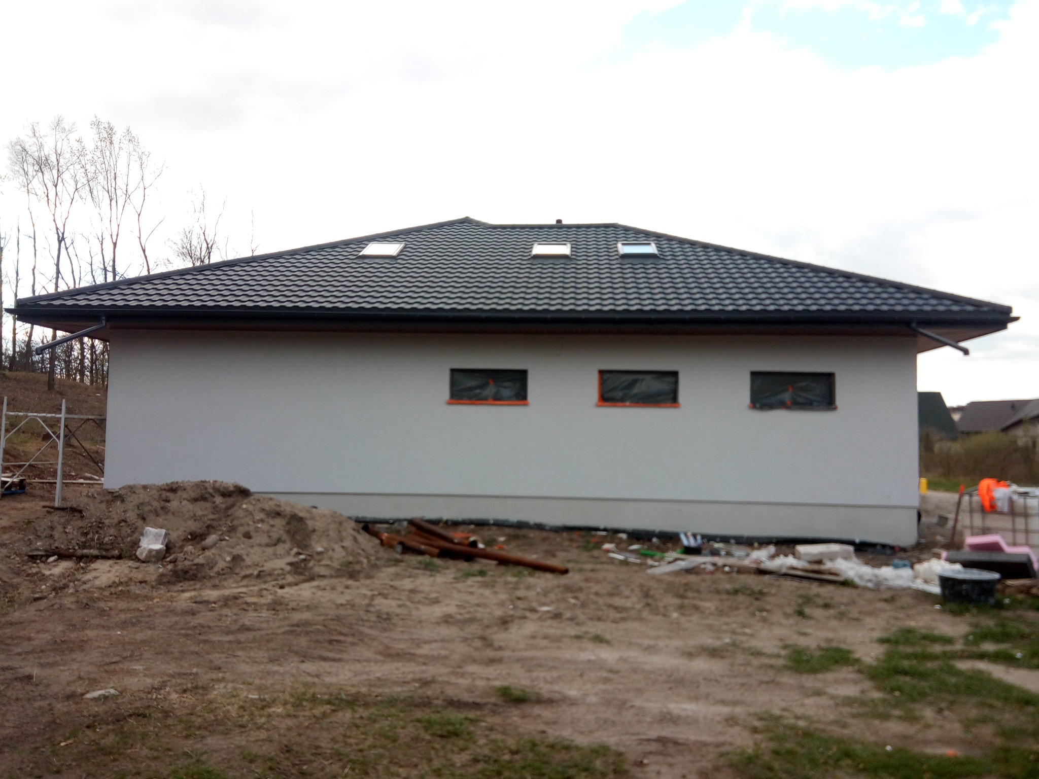 Niski, parterowy dom z ciemnym dachem i trzema oknami dachowymi. Ściany białe, okna zabezpieczone folią. Na pierwszym planie ziemia i materiały budowlane.