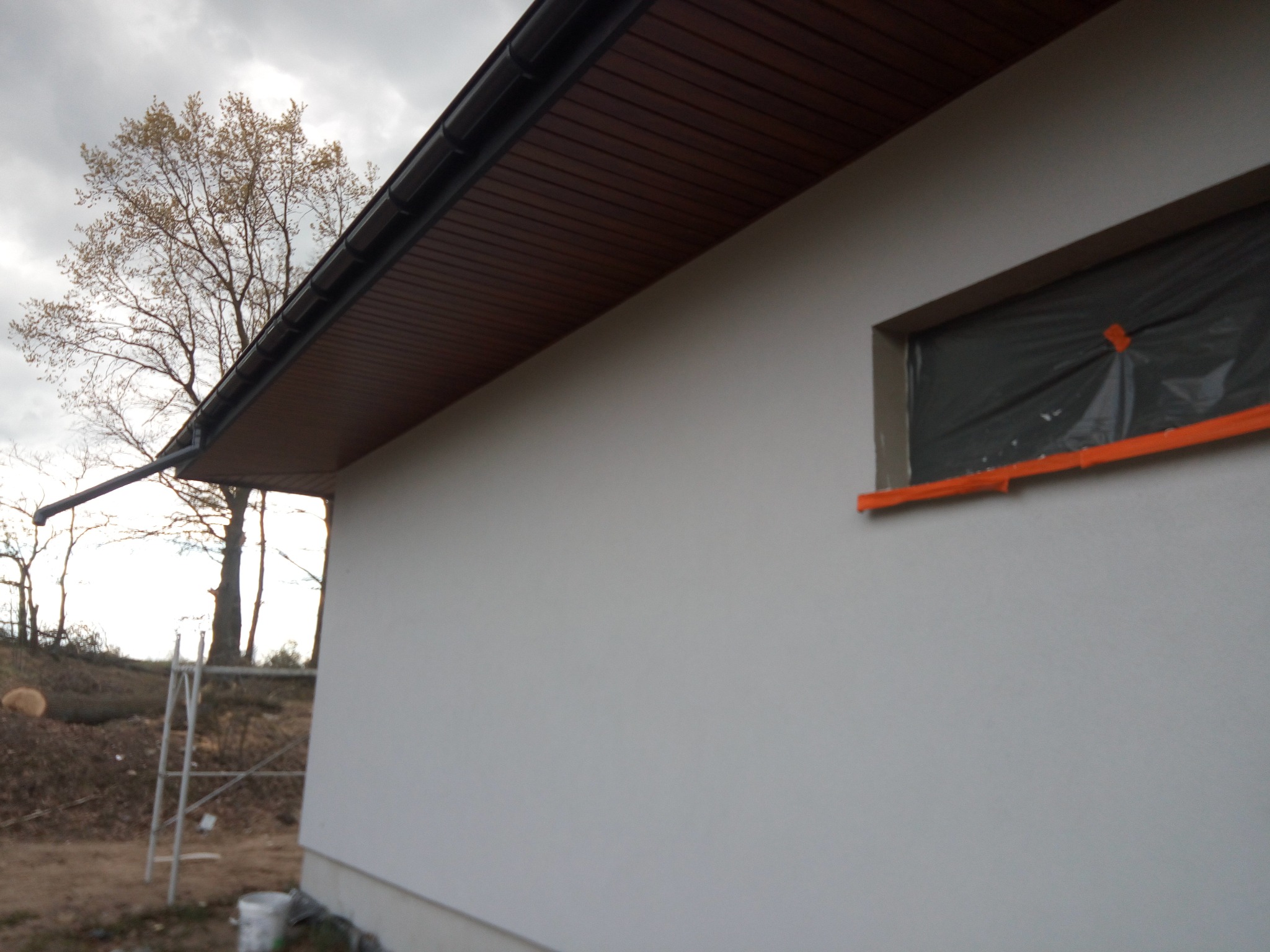 Fragment elewacji domu z zabezpieczonym oknem folią i widocznym fragmentem dachu z brązową podbitką. W tle drzewa i drabina.
