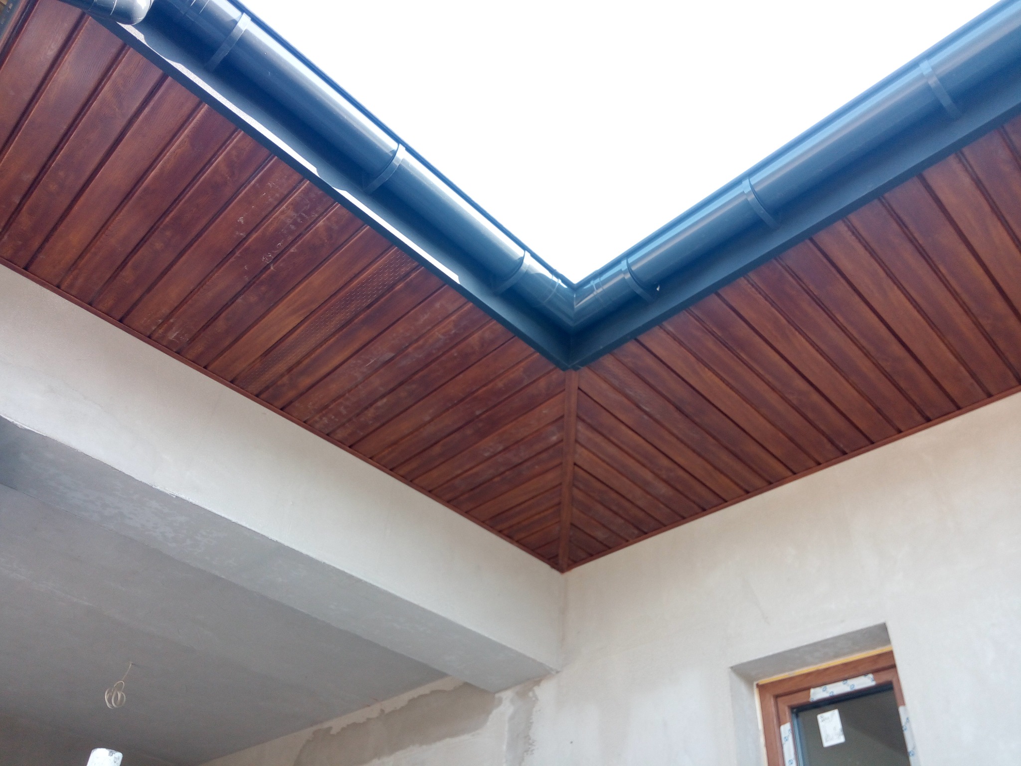 Drewniana podbitka dachowa w brązowym odcieniu, z zamontowaną rynną w kolorze grafitowym, widziana z dołu pod kątem. Widoczne fragmenty elewacji budynku.