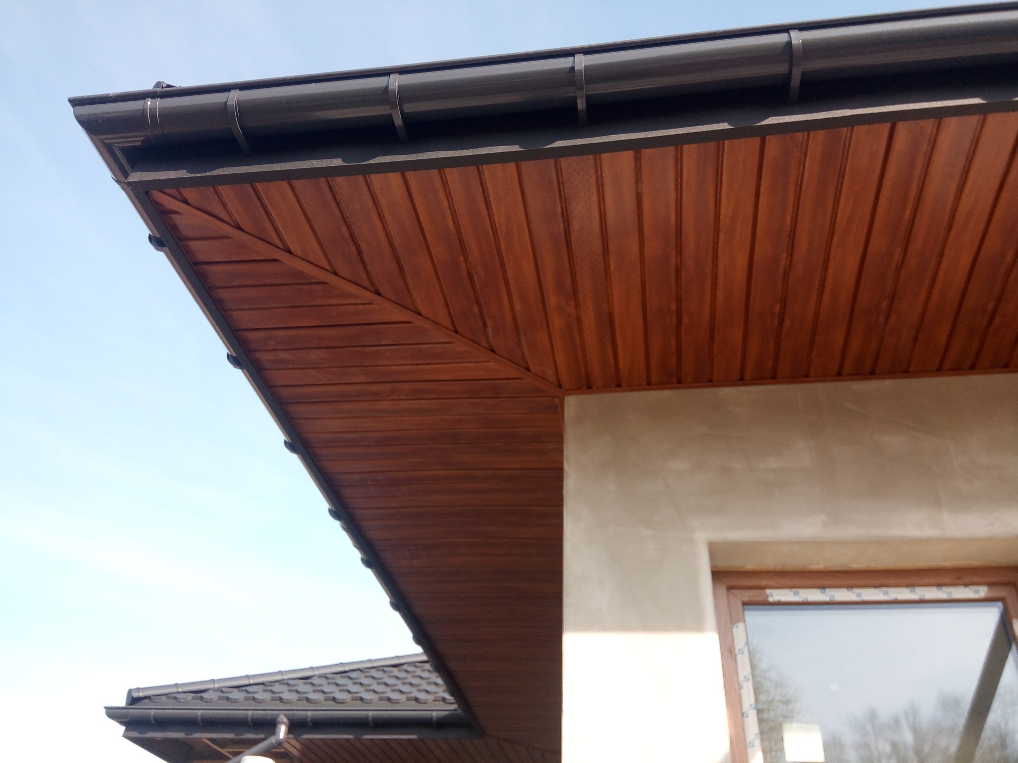 Drewniana podbitka dachowa z brązową rynną, widoczna z dołu pod kątem, fragment okna i elewacji budynku w tle, błękitne niebo.