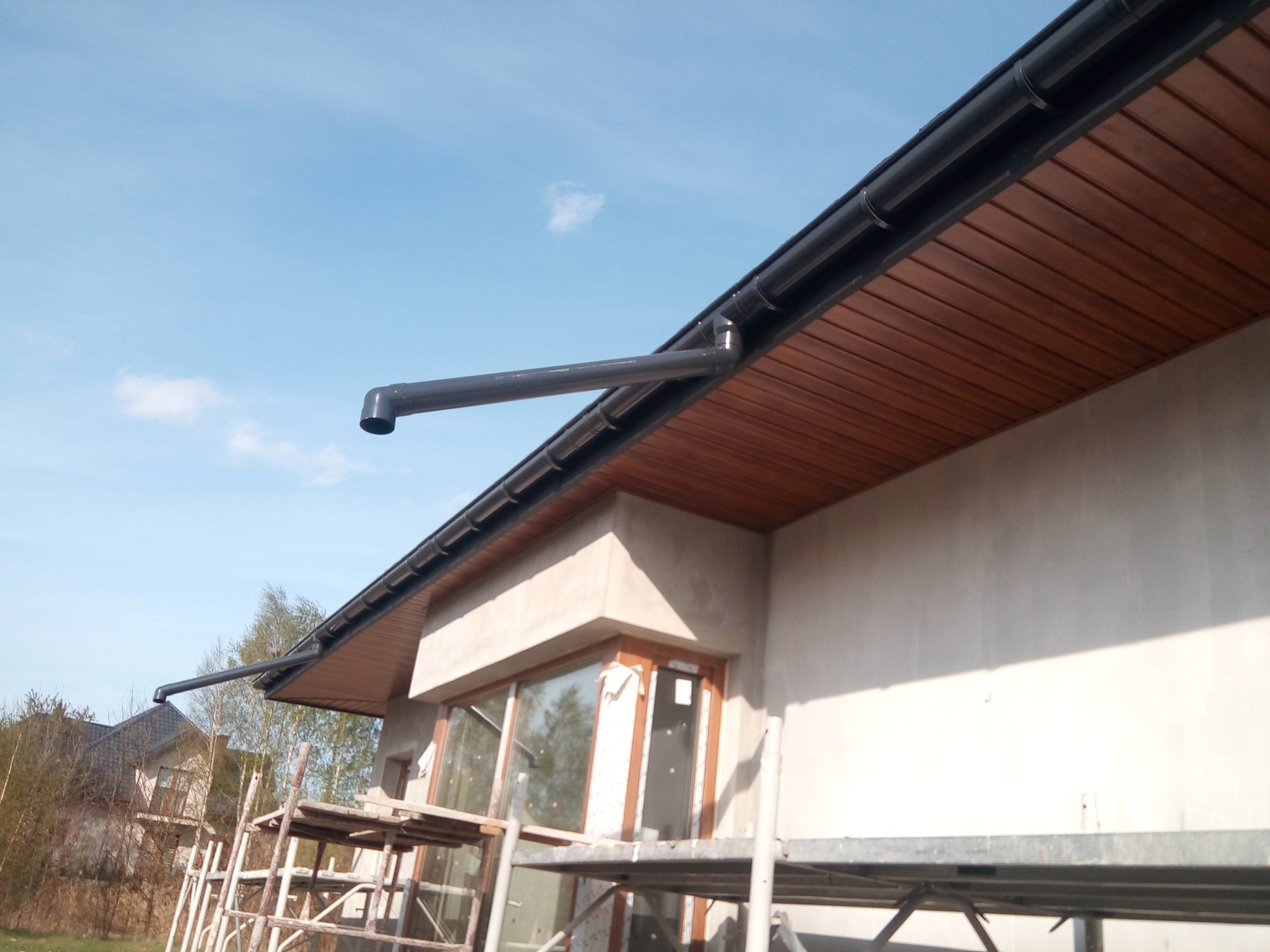 Surowy dom z drewnianą podbitką i czarnymi rynnami, widoczne rusztowania. Ujęcie z dołu, błękitne niebo w tle.