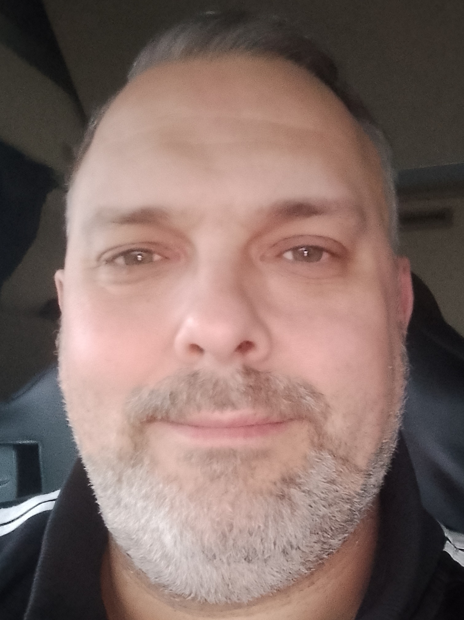 Portret mężczyzny z krótkimi, siwiejącymi włosami i lekkim zarostem, uśmiechającego się delikatnie. Ujęcie z bliska, z widocznym fragmentem ciemnej bluzy z białymi paskami.