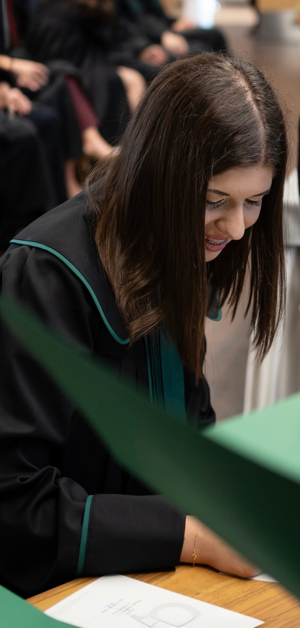 Młoda kobieta w todze prawniczej, pochylona nad dokumentami, być może podczas składania ślubowania. Widoczne detale: delikatna bransoletka, aparat ortodontyczny.