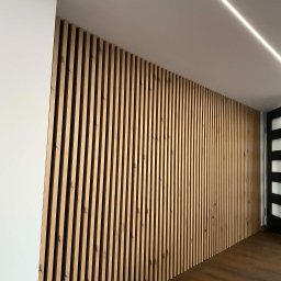 RemGol - Remonty i Tarasy Wentylowane - Drewniane lamele na ścianie, nowoczesny design wnętrza z oświetleniem LED na suficie i ciemną podłogą. Widoczny fragment białej ściany i czarne drzwi.