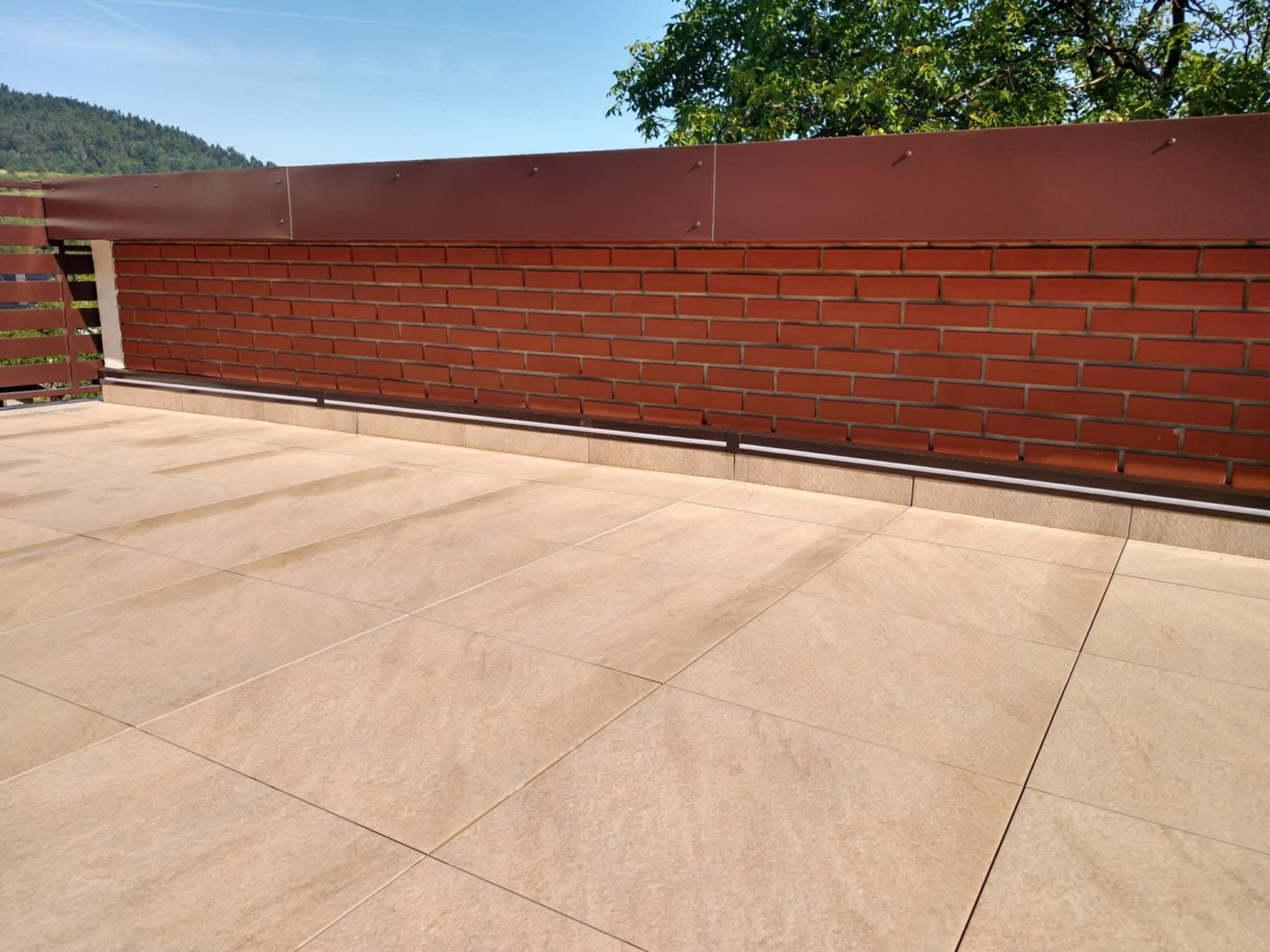 Wykończony balkon z beżowymi płytkami podłogowymi i ceglaną balustradą z brązowym panelem. Widok na zieleń w tle.