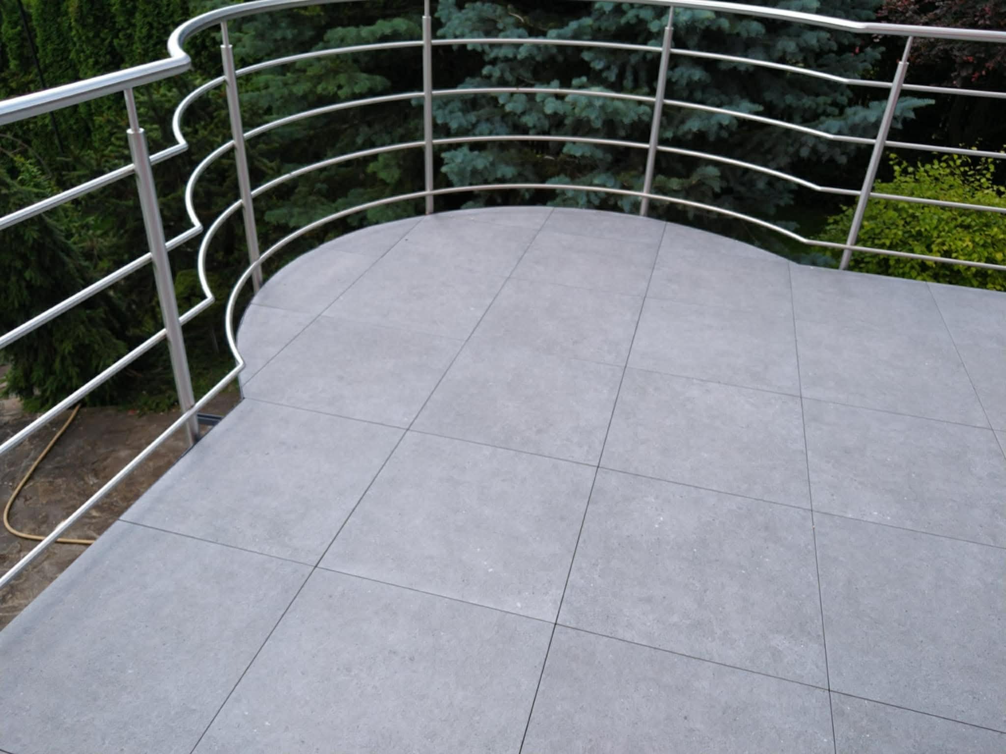 Wykończony balkon z szarymi płytkami i metalową balustradą na tle zieleni. Widok z góry, prezentujący estetyczne ułożenie płytek i nowoczesny design balustrady.