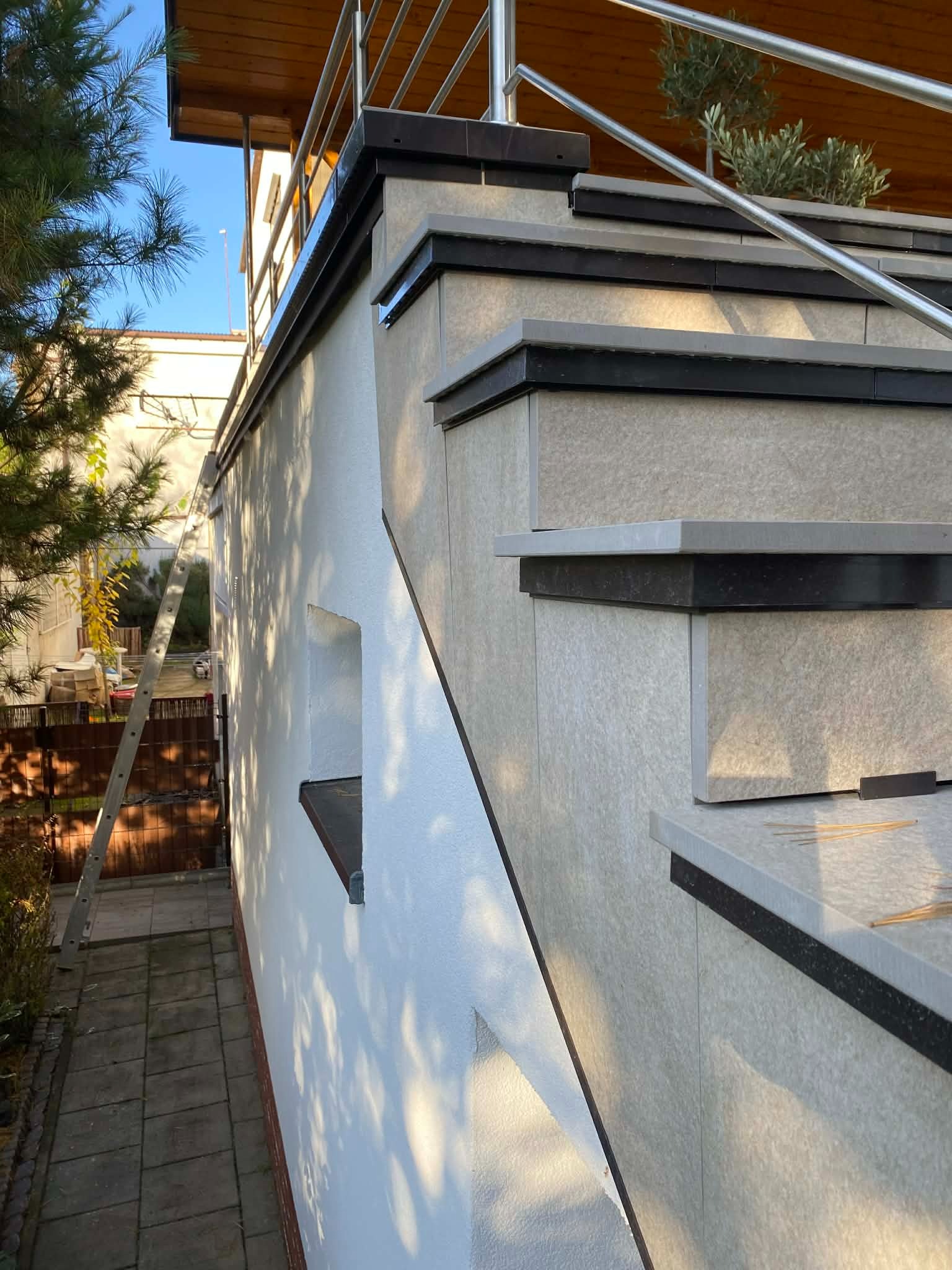 Wykończone schody zewnętrzne z jasnymi płytkami i ciemnymi detalami, widoczne z boku, z metalową barierką i fragmentem elewacji budynku w tle.