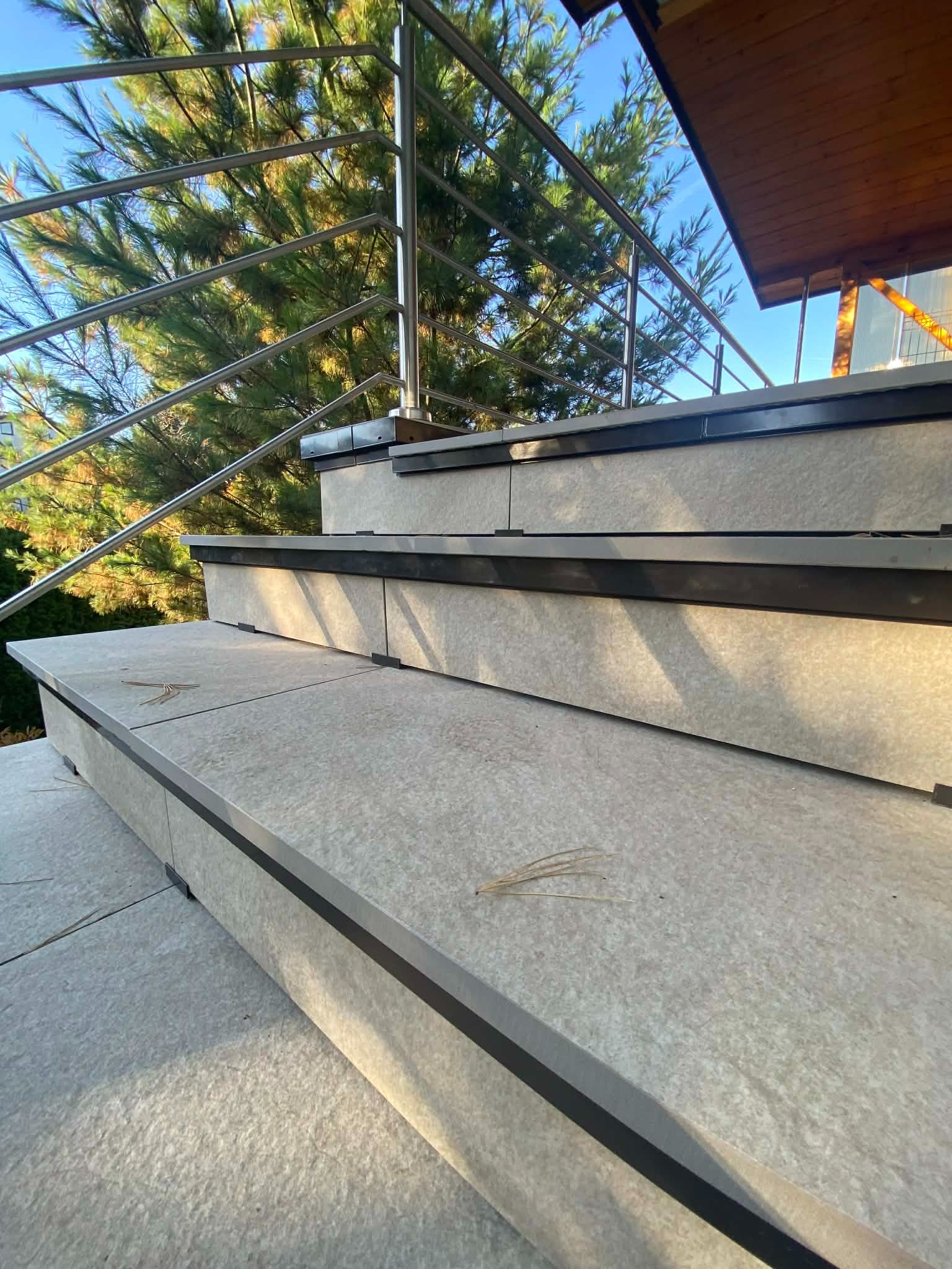 Wykończone schody zewnętrzne z szarych płyt z metalową balustradą na tle zieleni. Widoczne detale wykończenia i oświetlenie słoneczne.