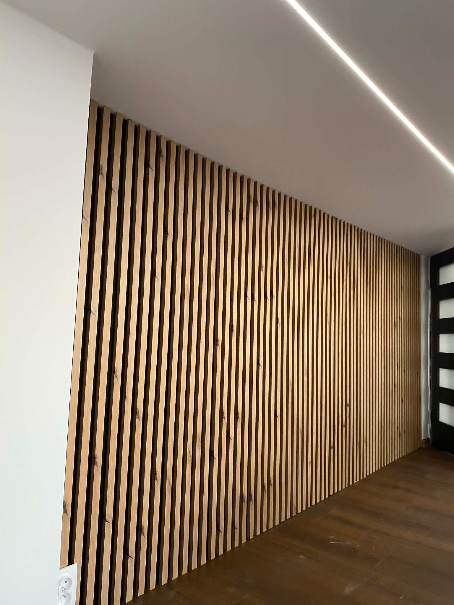Drewniane lamele na ścianie, nowoczesny design wnętrza z oświetleniem LED na suficie i ciemną podłogą. Widoczny fragment białej ściany i czarne drzwi.