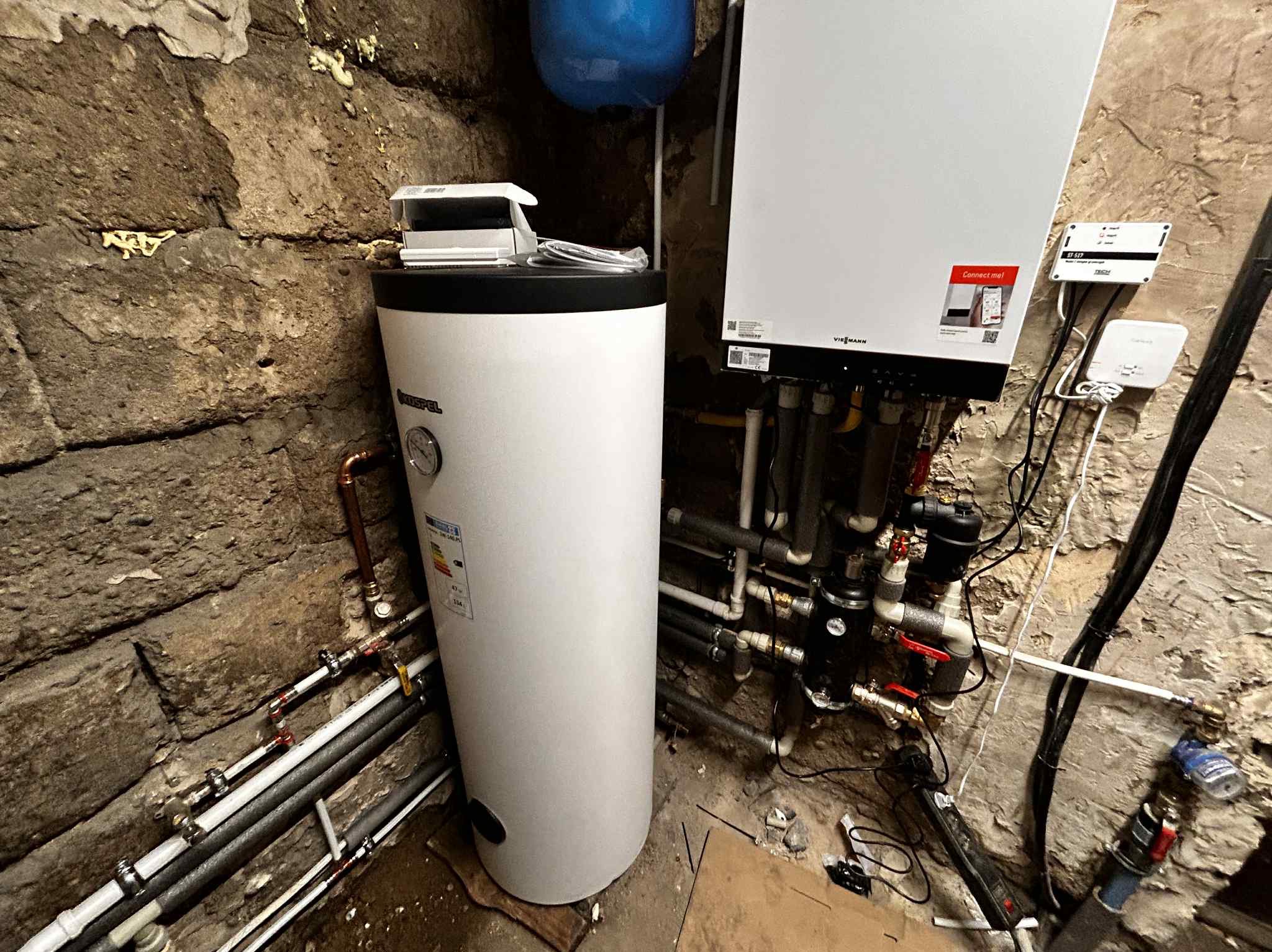 Biały zasobnik ciepłej wody Kospel i piec Viessmann w kotłowni z kamiennymi ścianami. Widoczne rury i instalacje.
