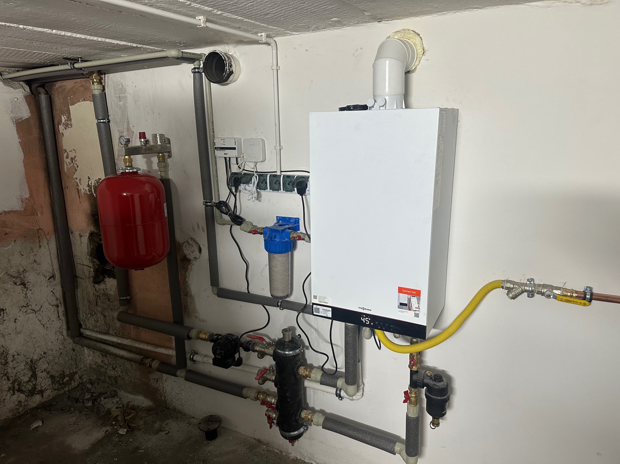 Biały piec grzewczy Viessmann z instalacją, rurami i czerwonym zbiornikiem wyrównawczym w piwnicy w Żurominie. Widoczne filtry i zawory.