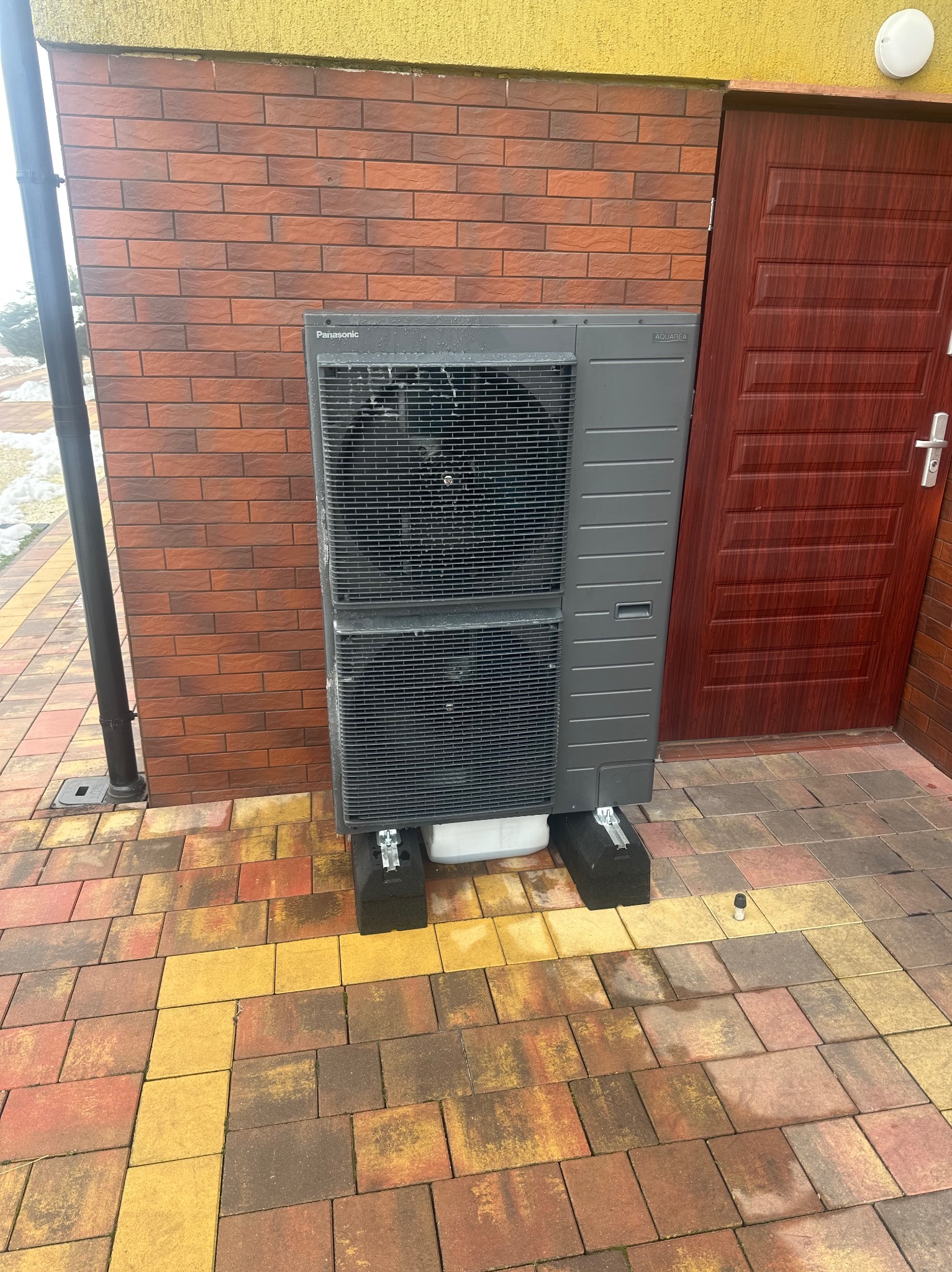 Szara pompa ciepła Panasonic Aquarea, zamontowana na zewnątrz budynku z ceglaną elewacją i brązowymi drzwiami, ustawiona na czarnych podstawach na kolorowej kostce brukowej.