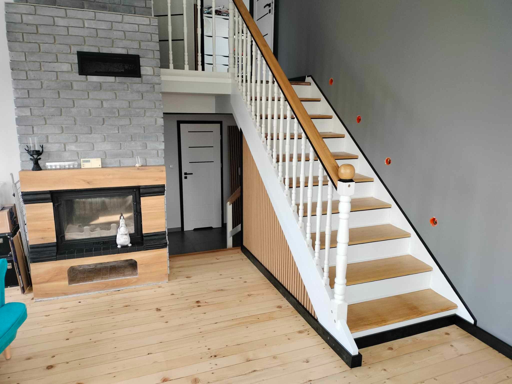 Jasne drewniane schody z białymi balustradami w nowoczesnym wnętrzu z kominkiem z szarej cegły i drewnianą podłogą. Szare ściany i białe drzwi w tle.