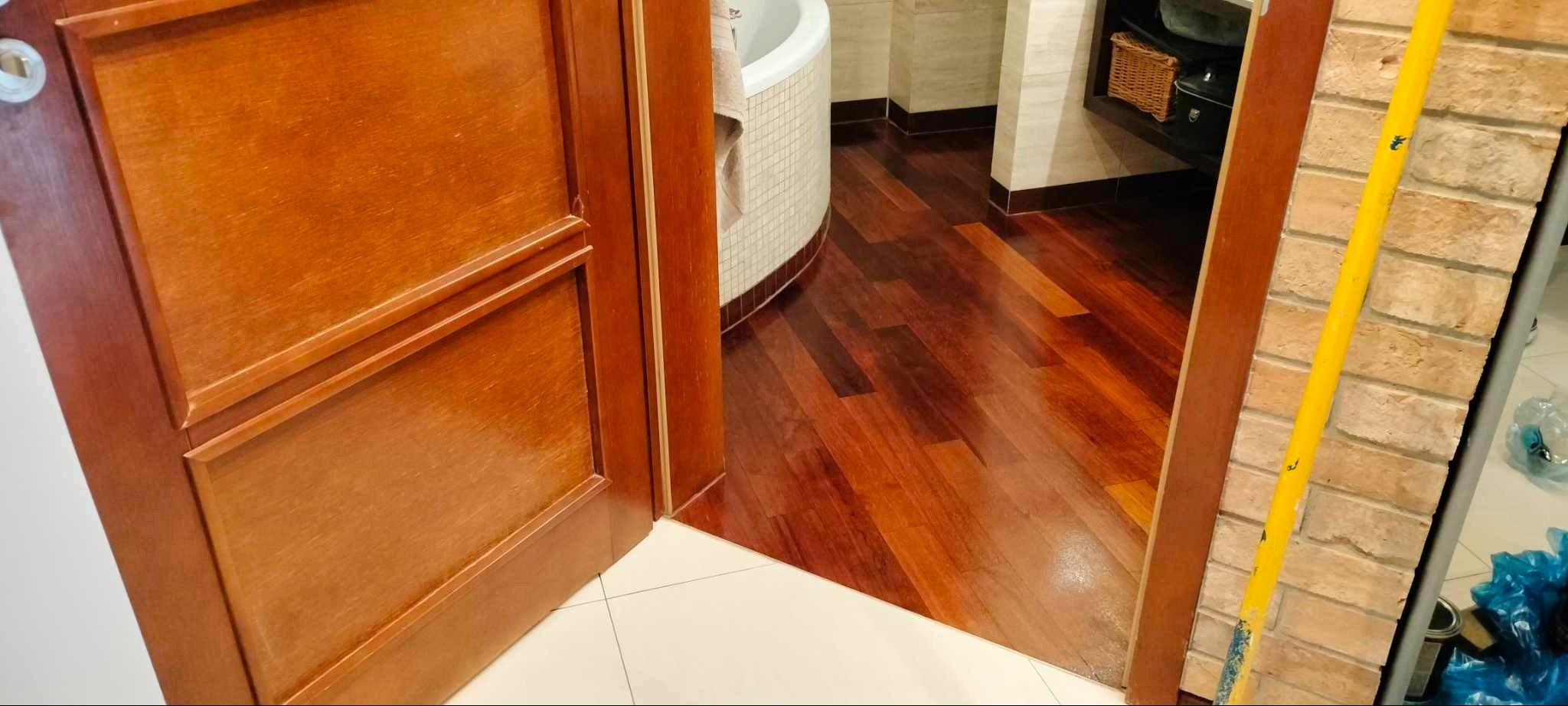 Otwarta drewniana rama drzwiowa z widokiem na błyszczący, ułożony parkiet w łazience z białą wanną i beżowymi płytkami ściennymi.